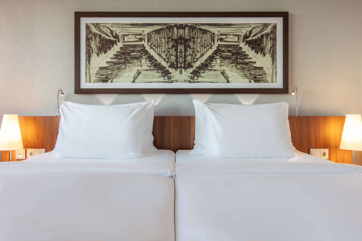 A double bed with white linens, two bedside lamps, and a framed artwork above the bed.