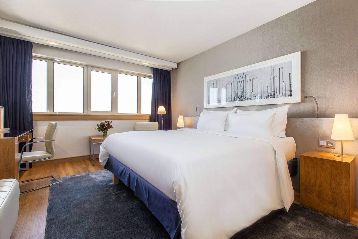 A hotel room with a large bed, two bedside tables with lamps, a chair, a desk, and a window with curtains.