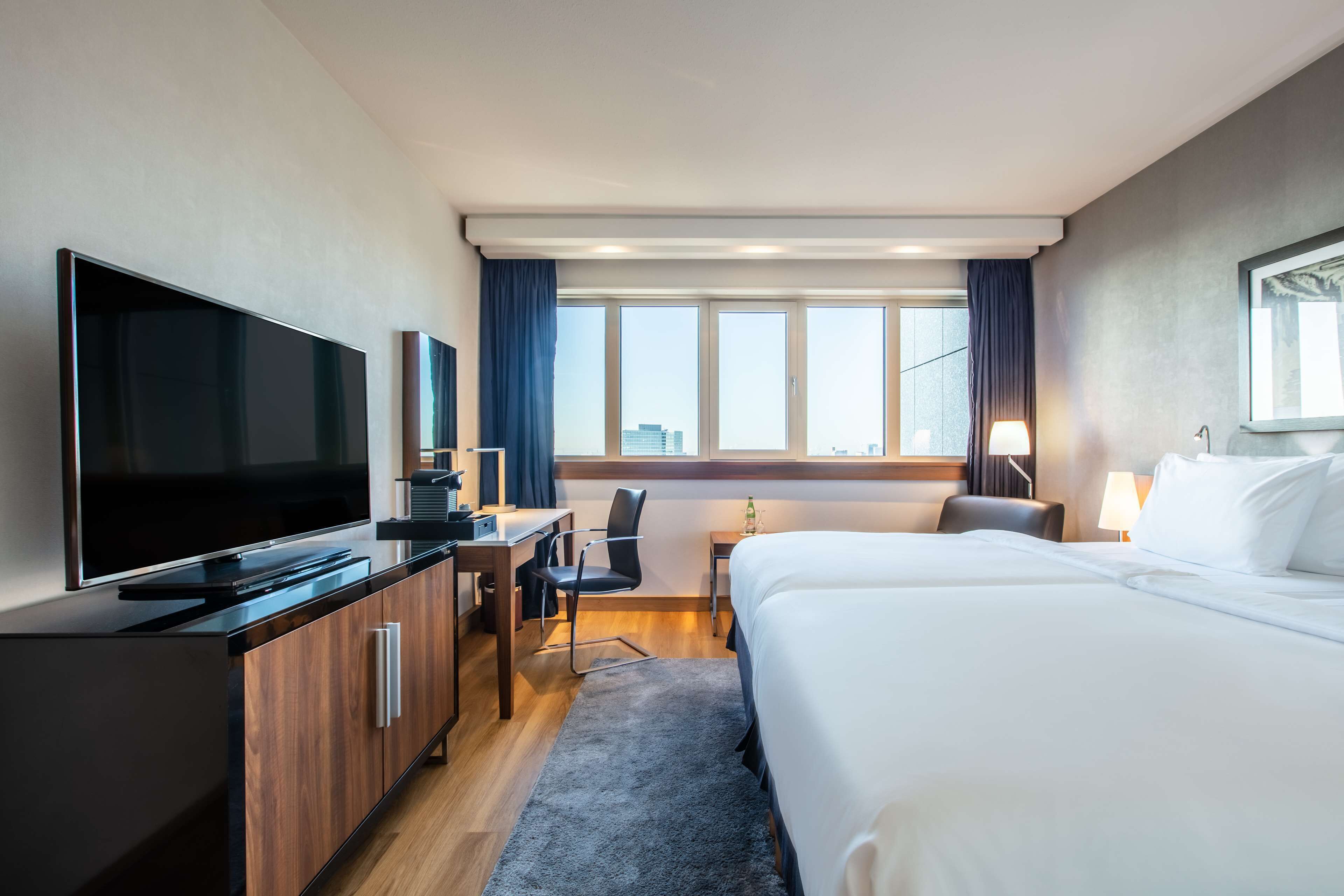 Ett hotellrum med en säng, ett skrivbord med en stol, en tv och ett fönster med gardiner.