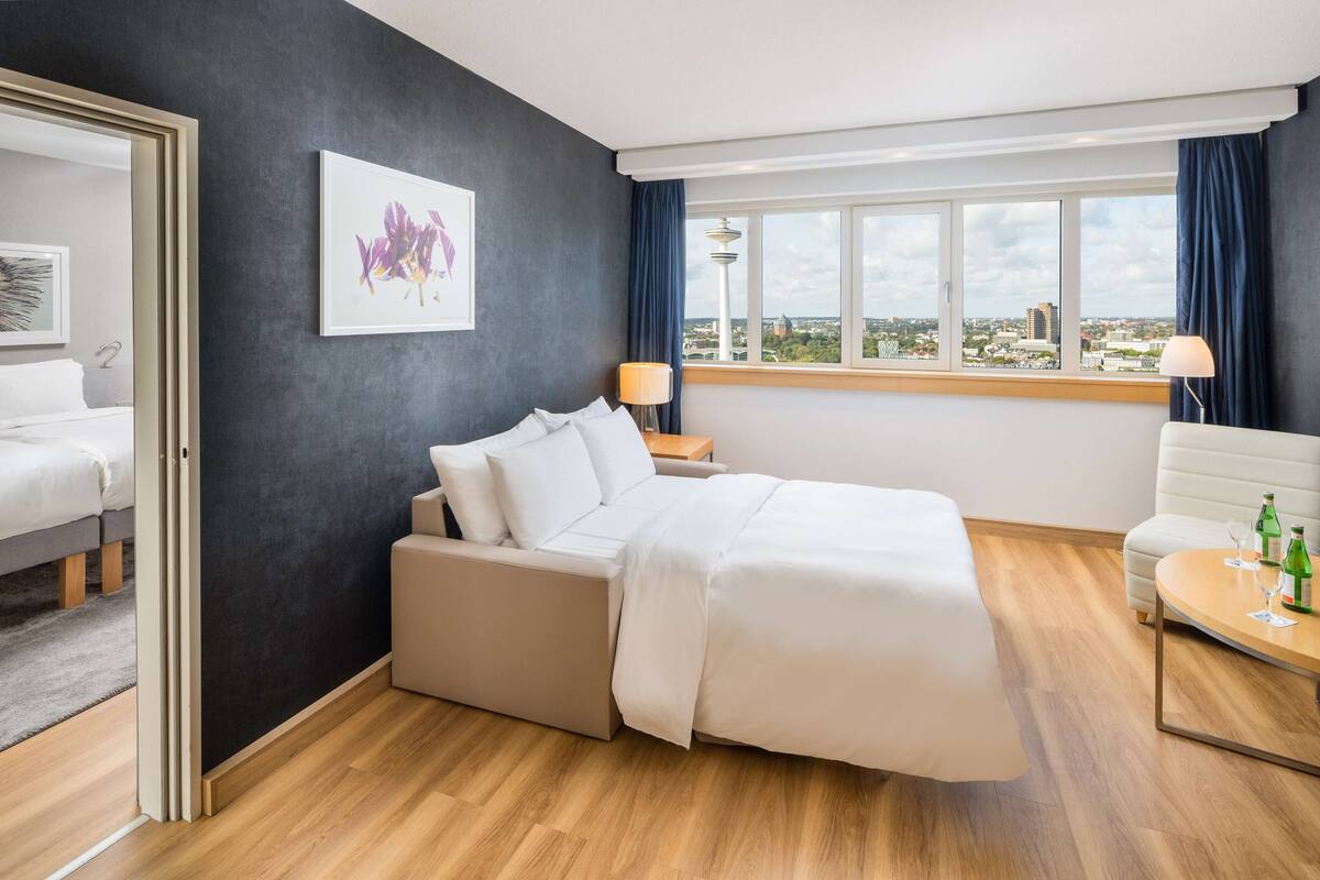 A modern hotel room with a bed, a sofa, a dining table, and a city view.