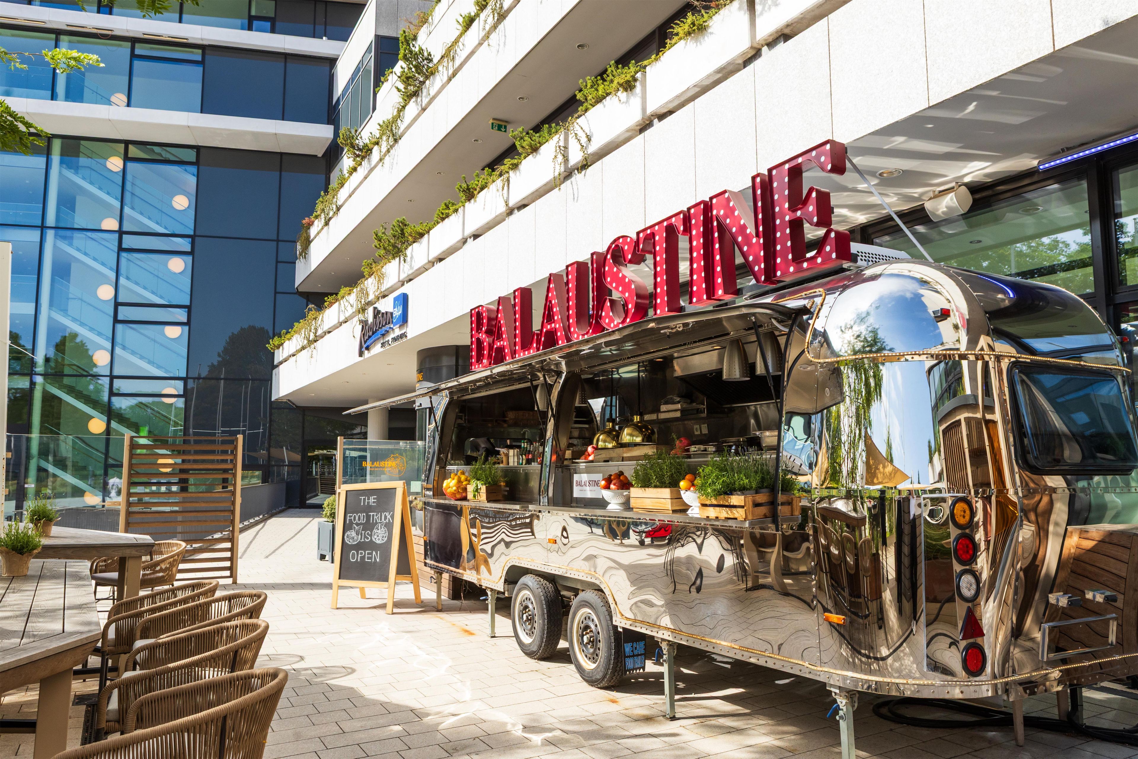 Ein Food-Truck mit dem Schriftzug 'BALASTINE', parkt vor einem Gebäude mit Glasfassade und Außenplätzen.