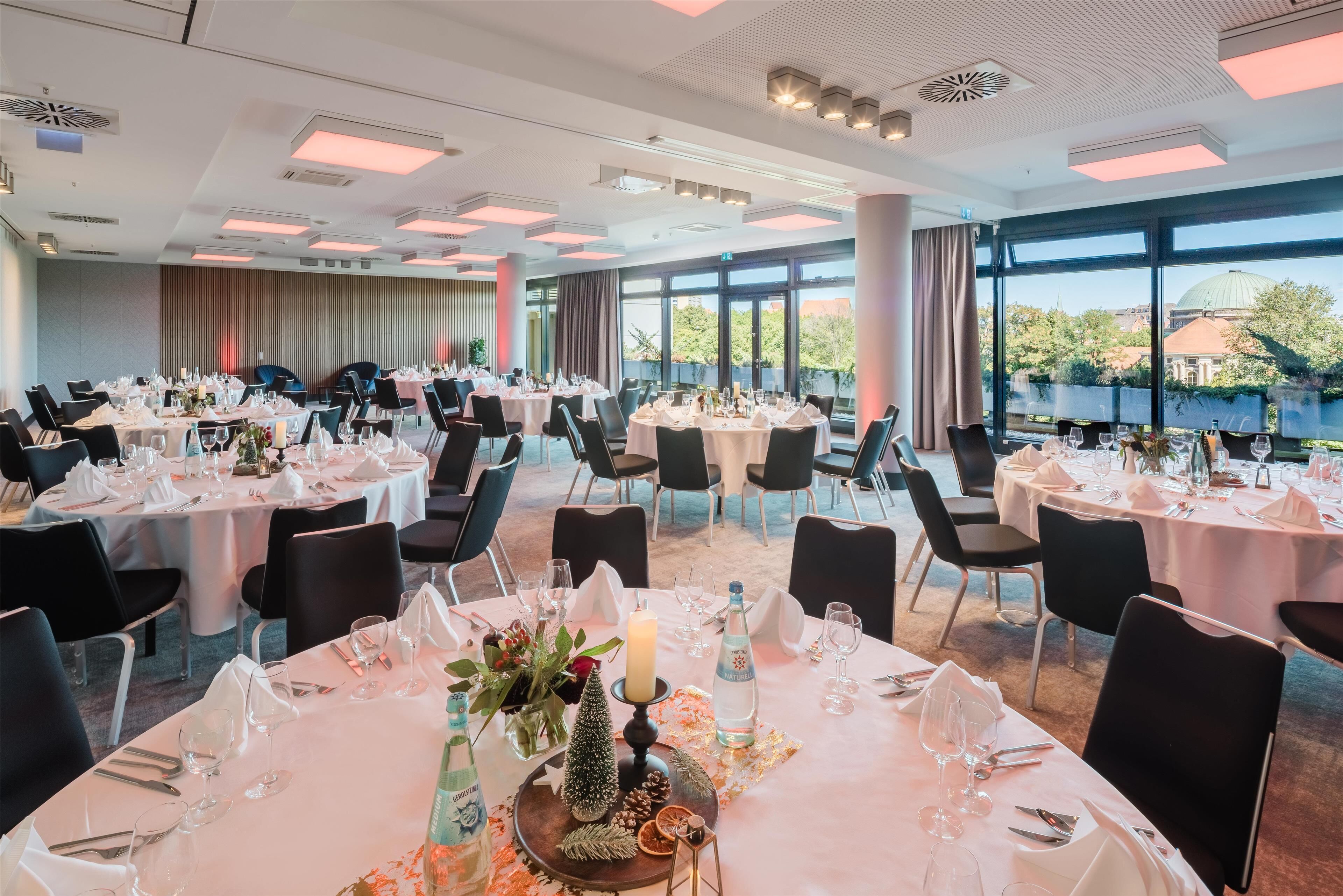 A banquet hall with round tables set for dining, large windows offering a view of the outdoors, and a modern ceiling design.