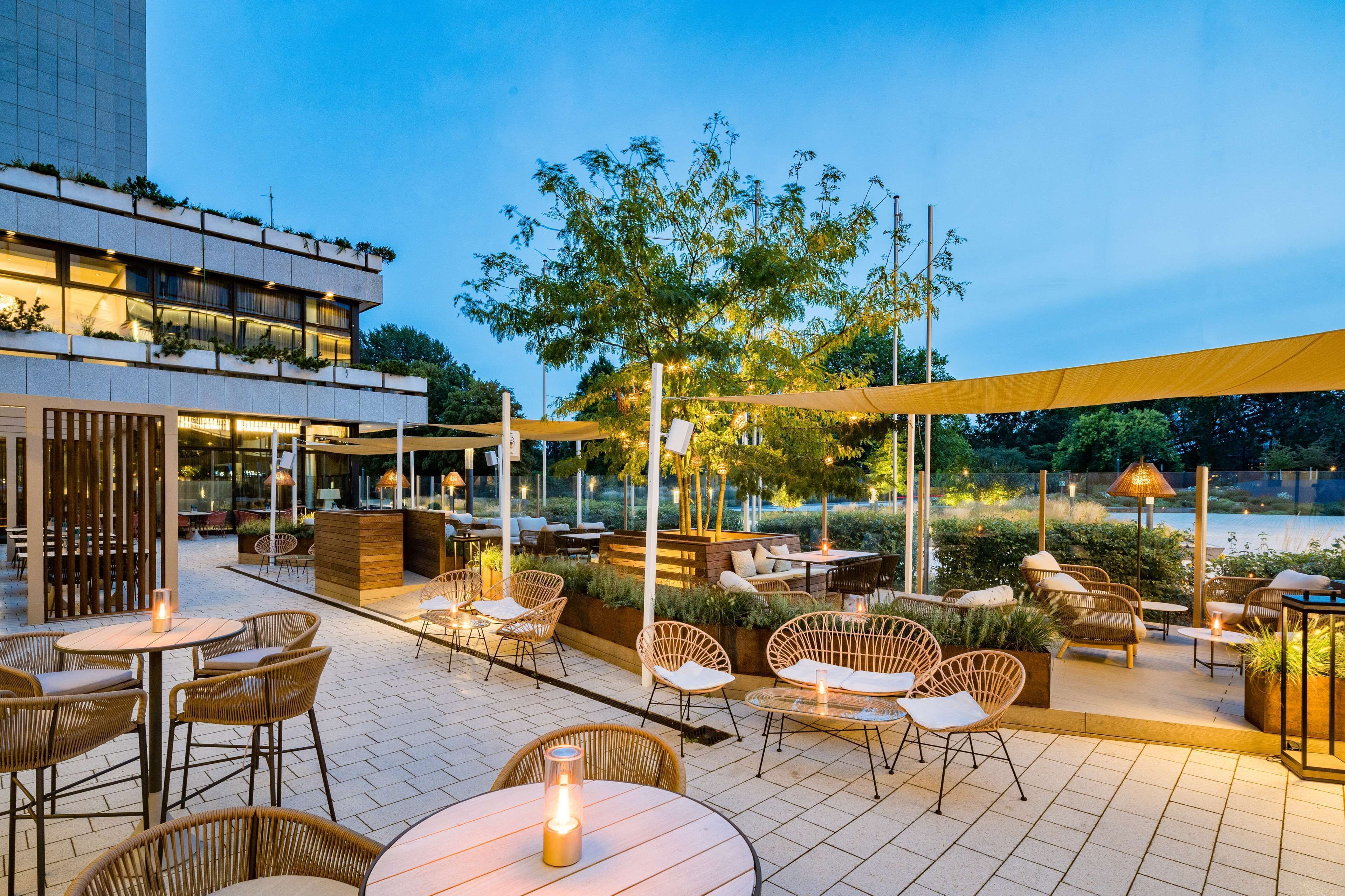 An outdoor patio area with wicker furniture, tables, and a large tree providing shade.