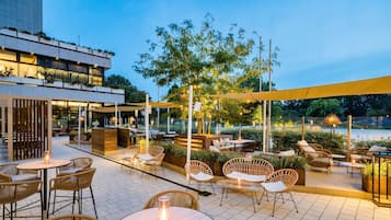 Un patio al aire libre con mobiliario de mimbre, mesas y un árbol grande que da sombra.