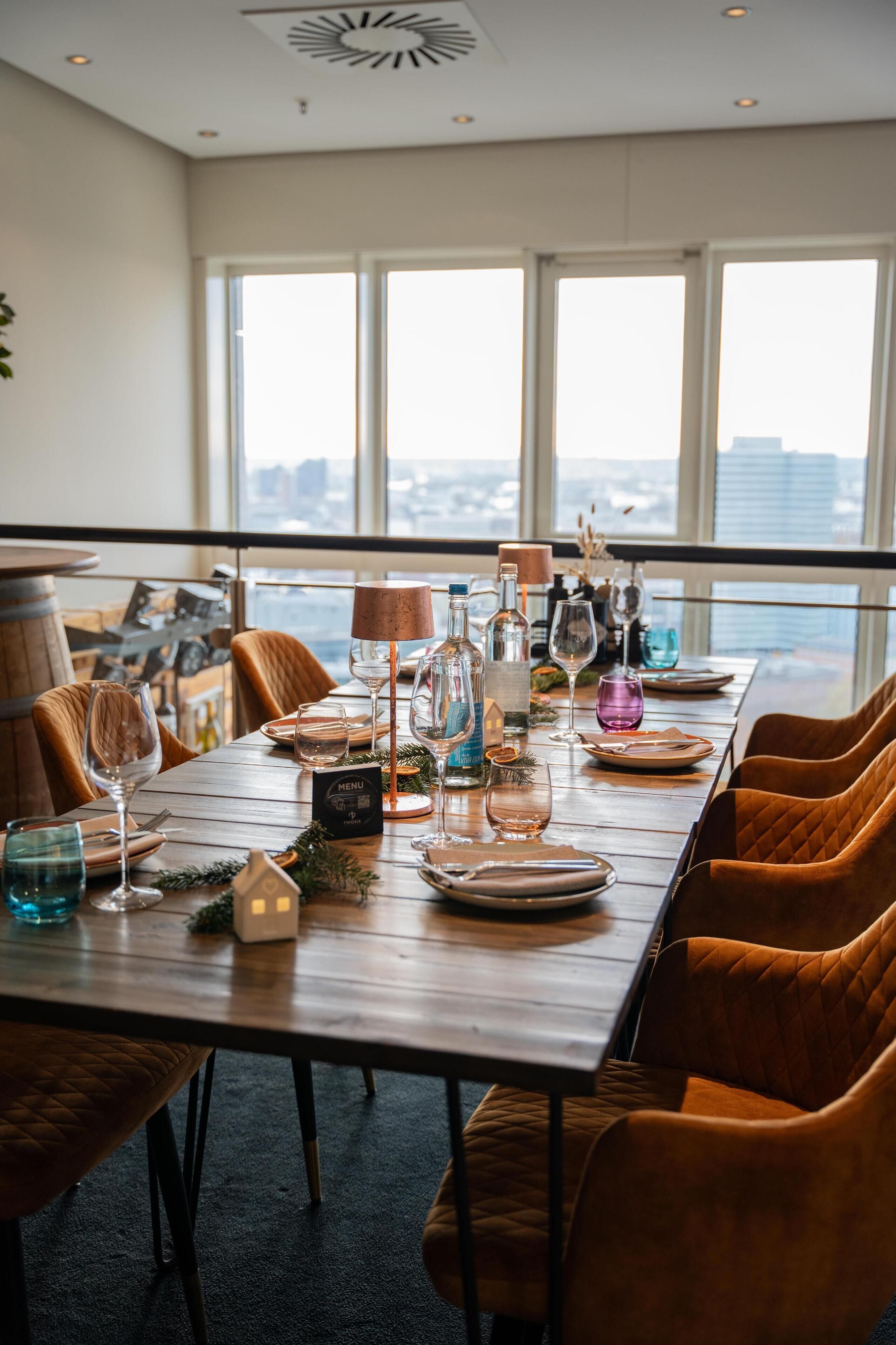 A dining table set with wine glasses, bottles, and plates, overlooking a cityscape through large windows.