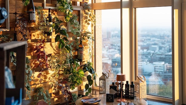 Ein gemütliches Restaurant mit einer Holzwand, die mit Pflanzen und Weinflaschen dekoriert ist, einem runden Tisch mit Stühlen und Blick auf die Stadt durch große Fenster.