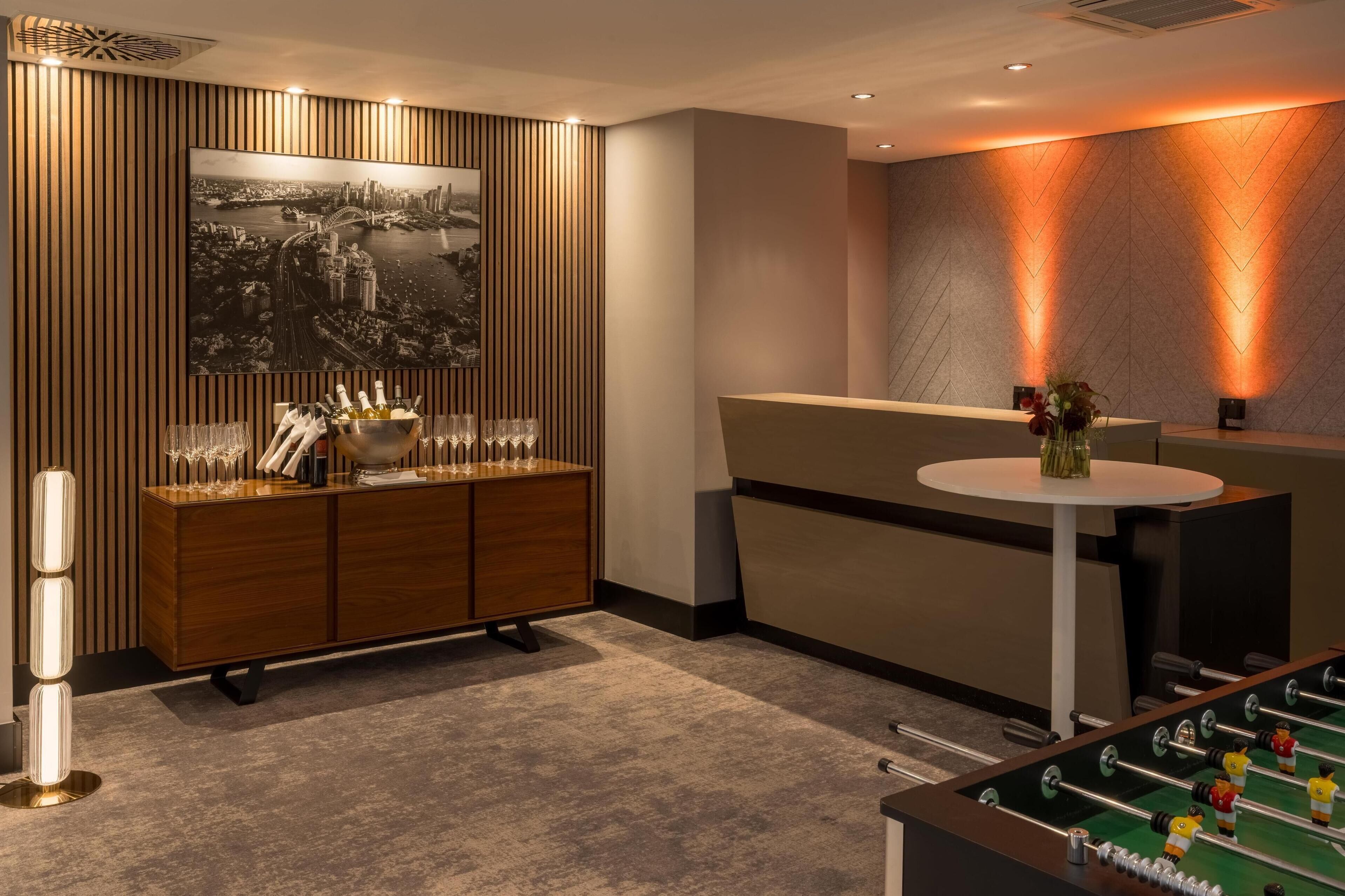 A modern hotel lobby with a wooden panel wall, a large cityscape painting, a reception desk, a foosball table, and a bar area with bottles and glasses.
