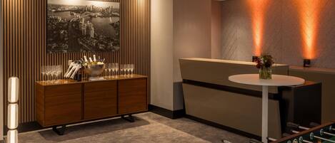 A modern hotel lobby with a wooden panel wall, a large cityscape painting, a reception desk, a foosball table, and a bar area with bottles and glasses.