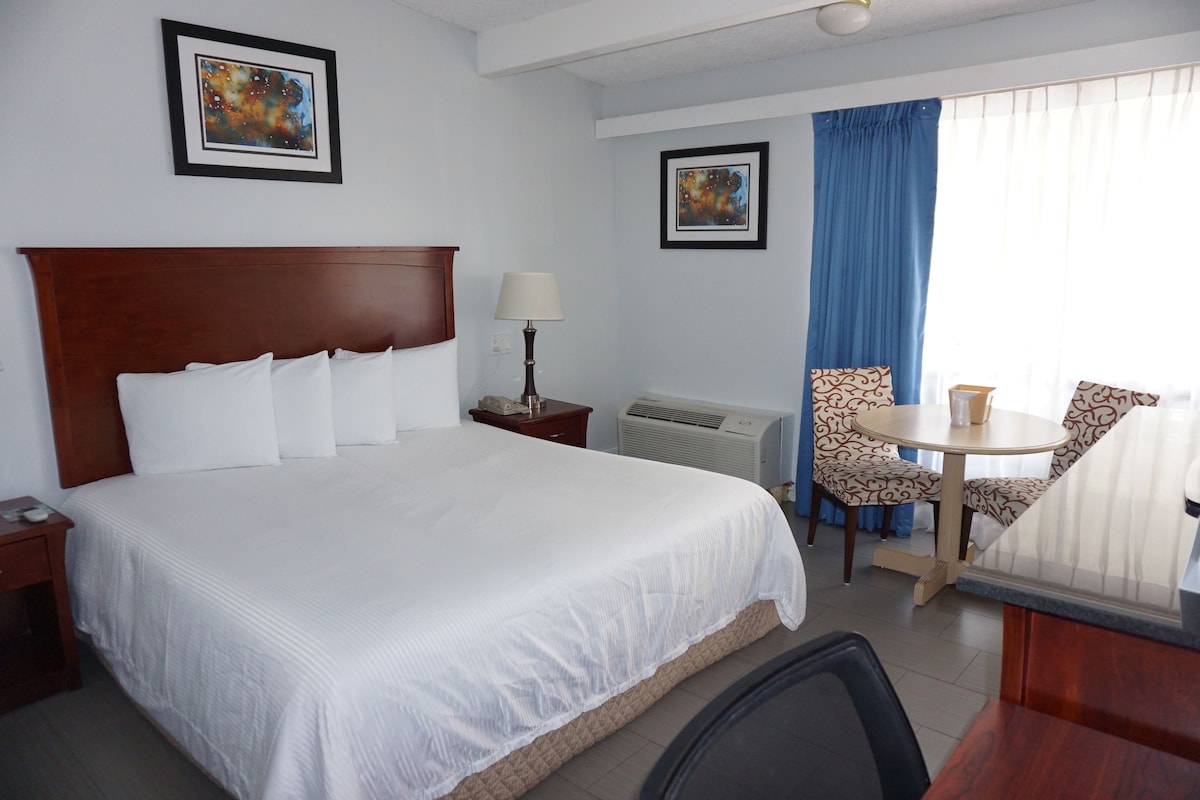 A hotel room with a large bed, a bedside table with a lamp, a small round table with a chair, and two framed artworks on the walls.