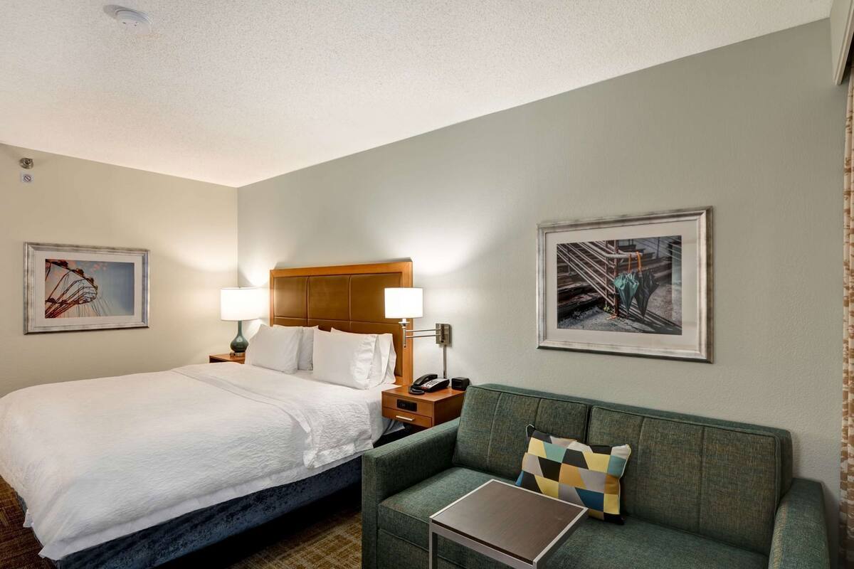 A hotel room with a bed, a green sofa, two bedside tables with lamps, and two framed pictures on the walls.