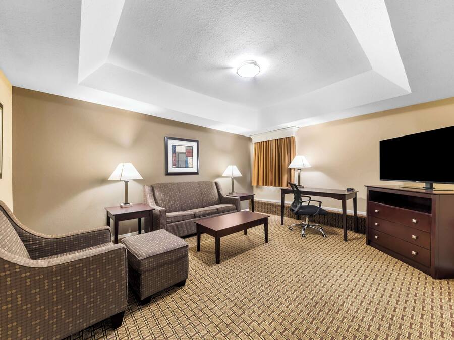 Quarto de hotel com sofá, poltrona, mesa de centro, suporte para TV e piso coberto de carpete.