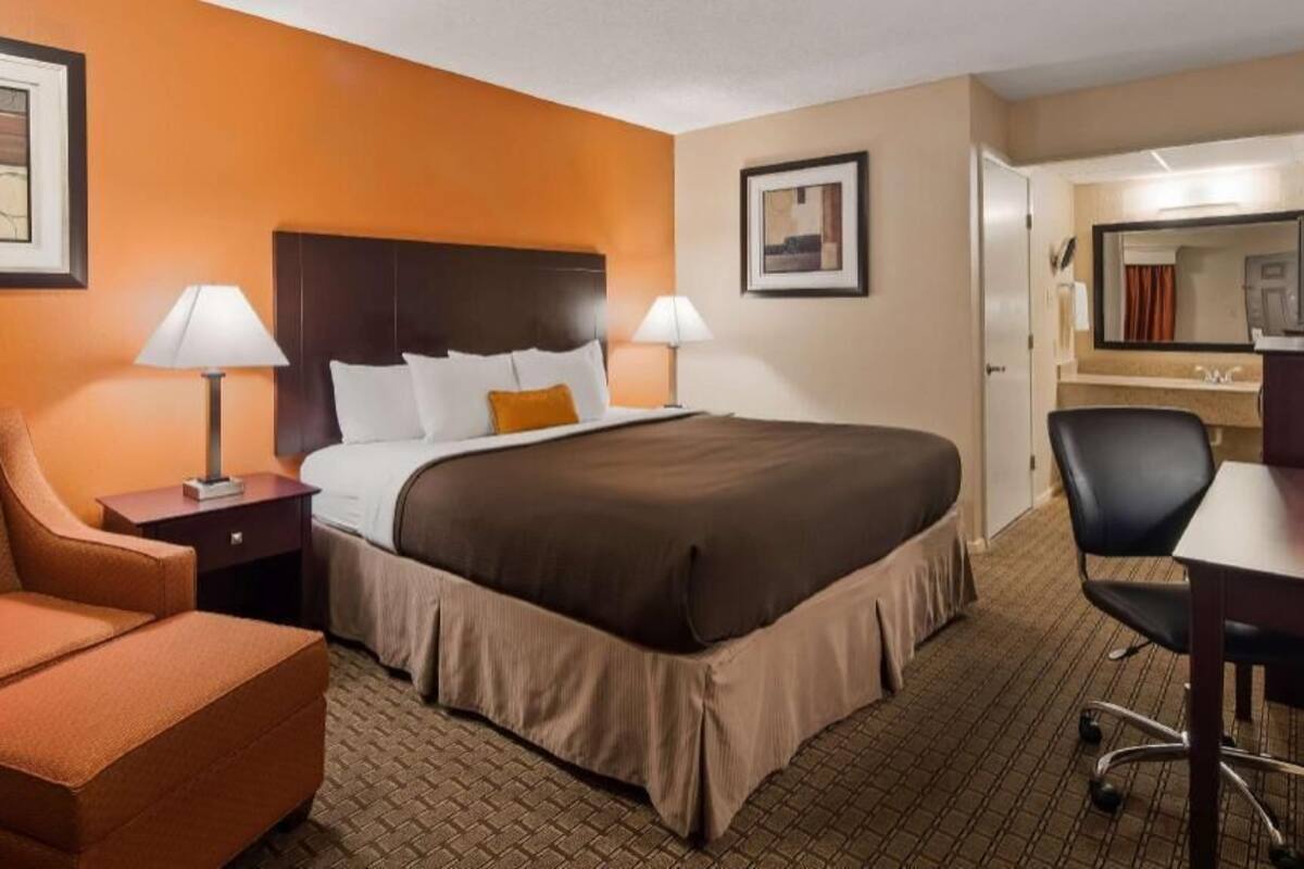 Ein Hotelzimmer mit einem Bett, zwei Nachttischlampen, einem Schreibtisch mit Stuhl, einem kleinen orangefarbenen Sessel und einem durch eine offene Tür einsehbaren Badezimmer.
