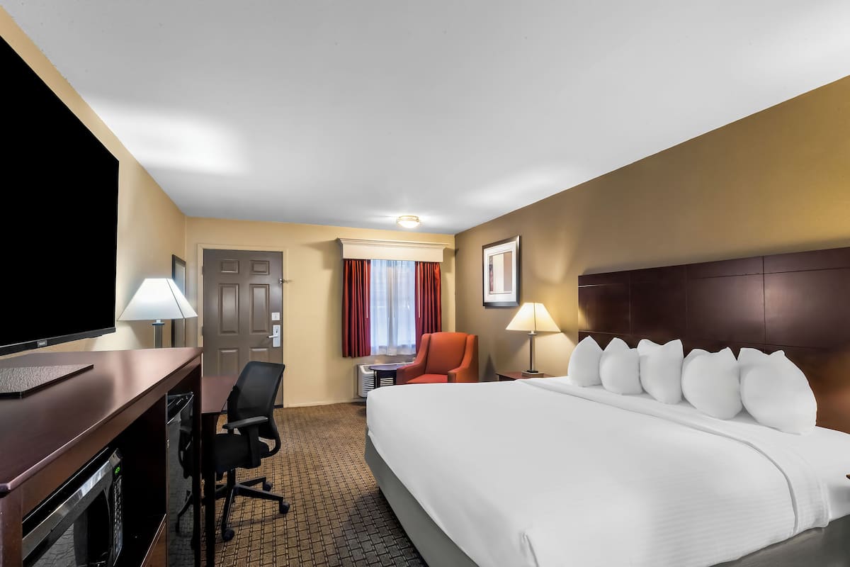 Ein Hotelzimmer mit einem großen Bett, einem Schreibtisch mit Fernseher, einem Sessel und einem kleinen Tisch mit Lampe.