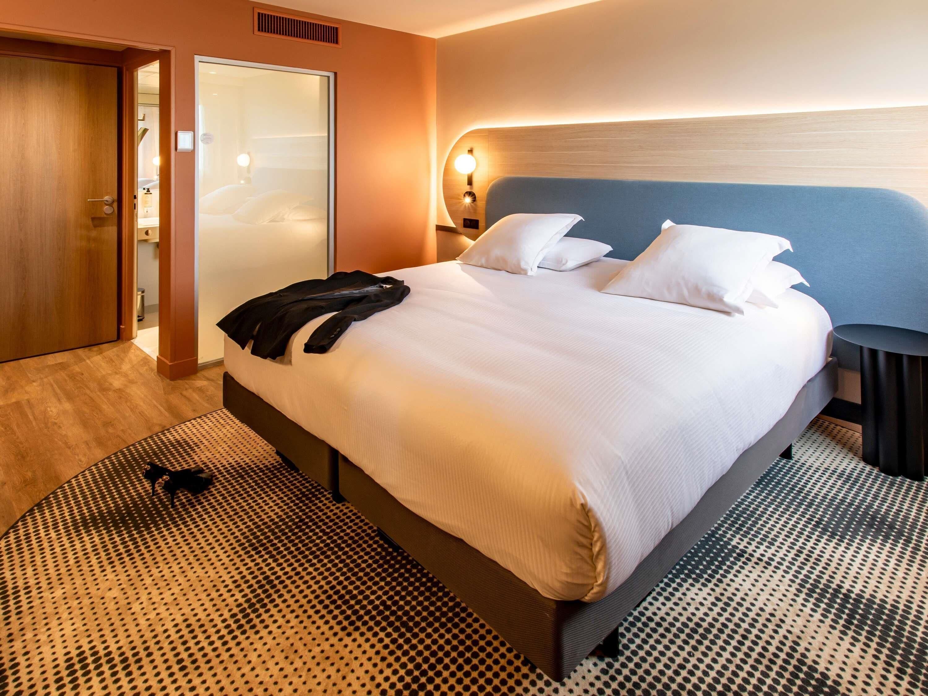 A hotel room with a bed, a nightstand, a wall-mounted lamp, a wardrobe, and a patterned rug.