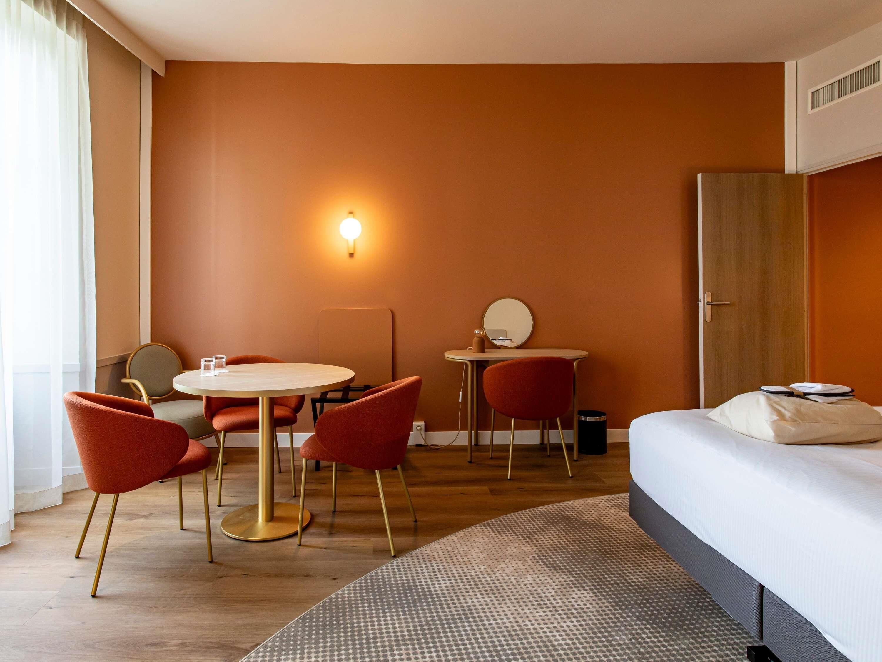 A hotel room with a bed, a round table with chairs, a small desk with a mirror, and a wall-mounted light.