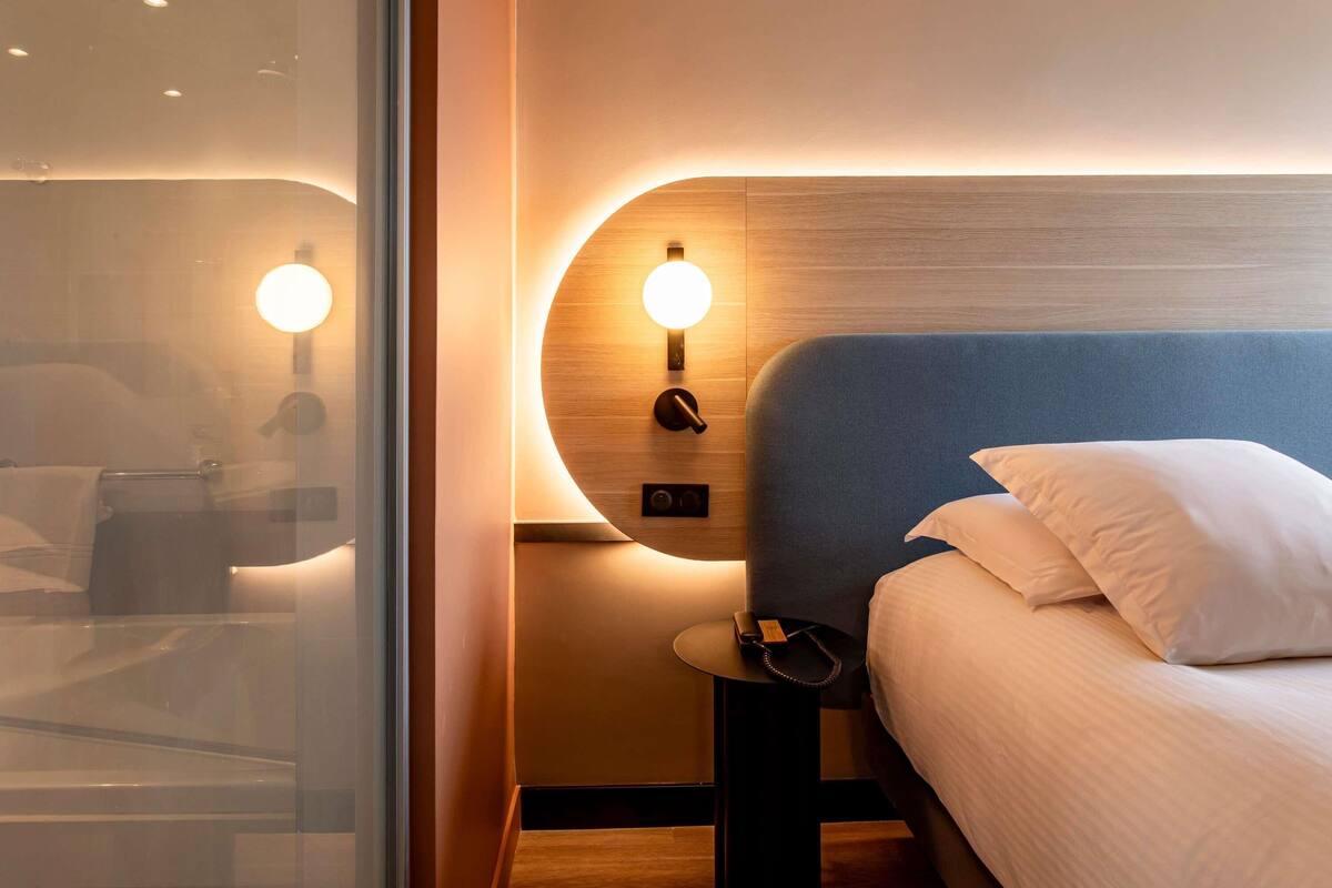 A modern hotel room with a curved headboard, bedside table, and a large bed with white bedding.