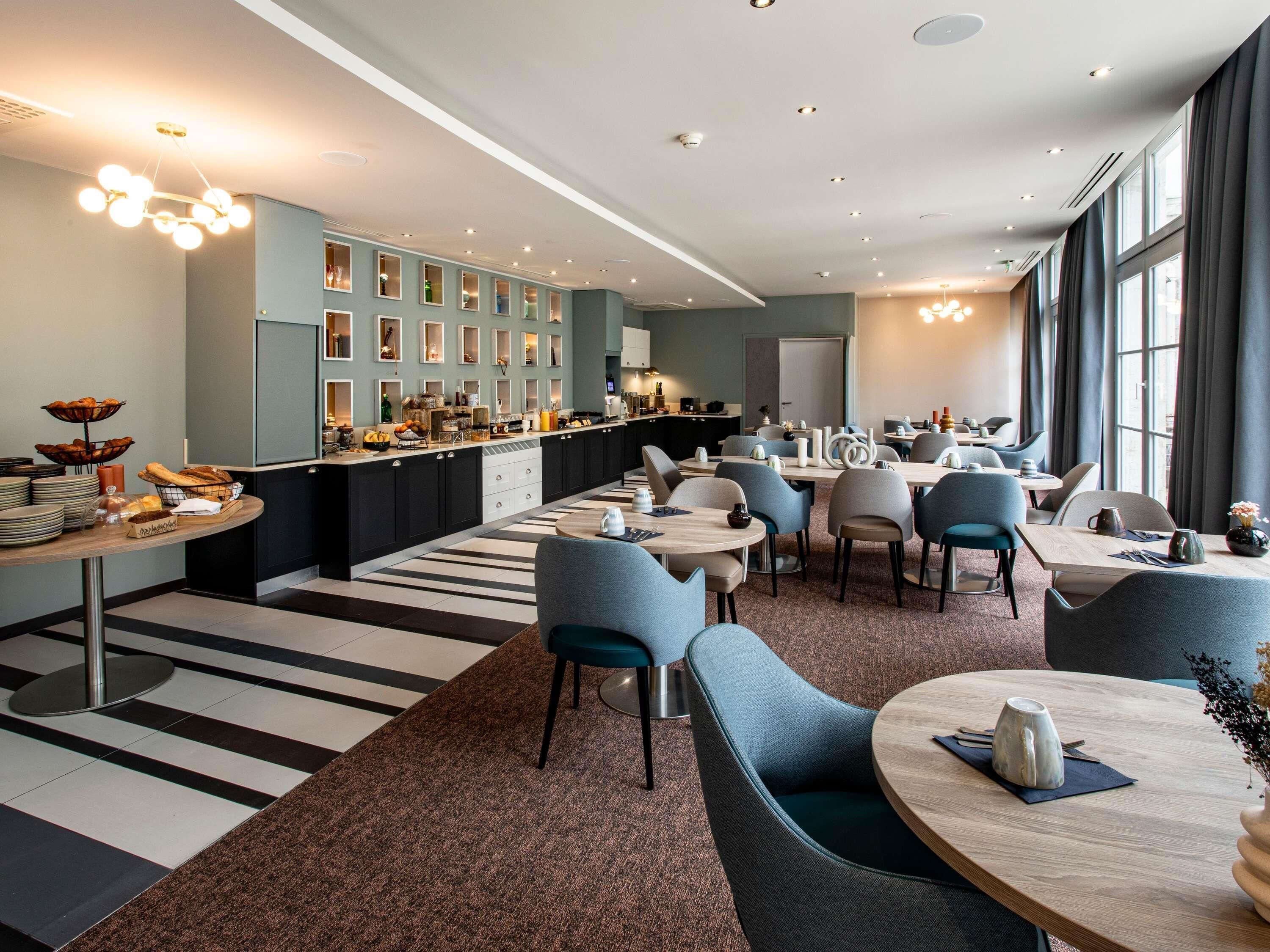Un espace de restauration moderne dans un hôtel, avec un bar central, de nombreux espaces de détente et une cuisine située à l’arrière-plan.
