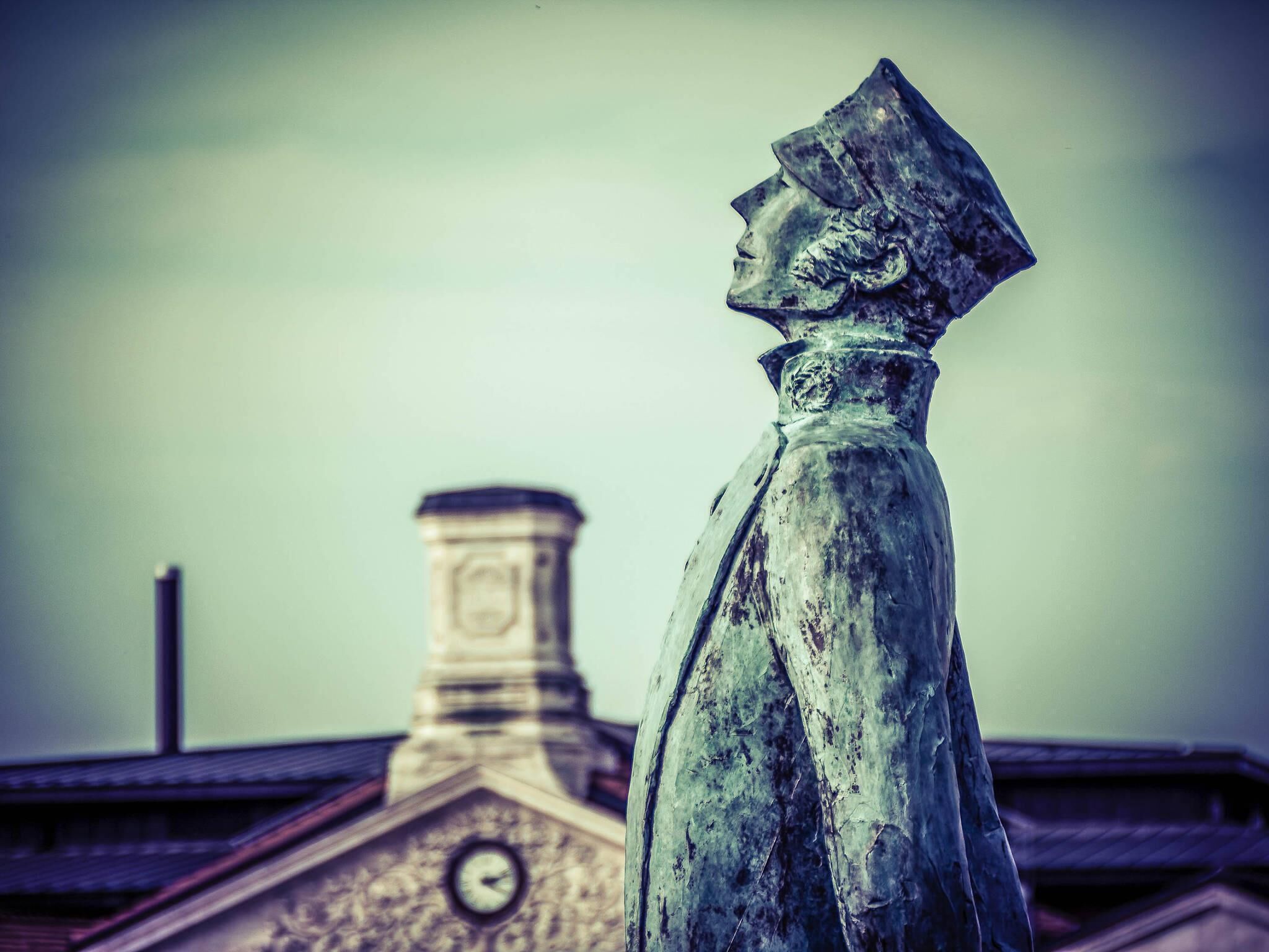 A statue of a person with a hat, a clock tower, and a chimney in the background.