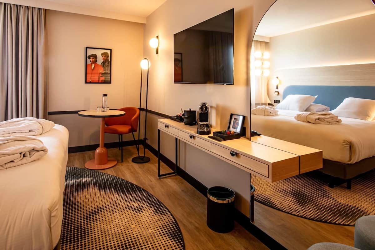 Une chambre d’hôtel comprenant un lit, un bureau avec une chaise, une petite table, une poubelle et un grand miroir.