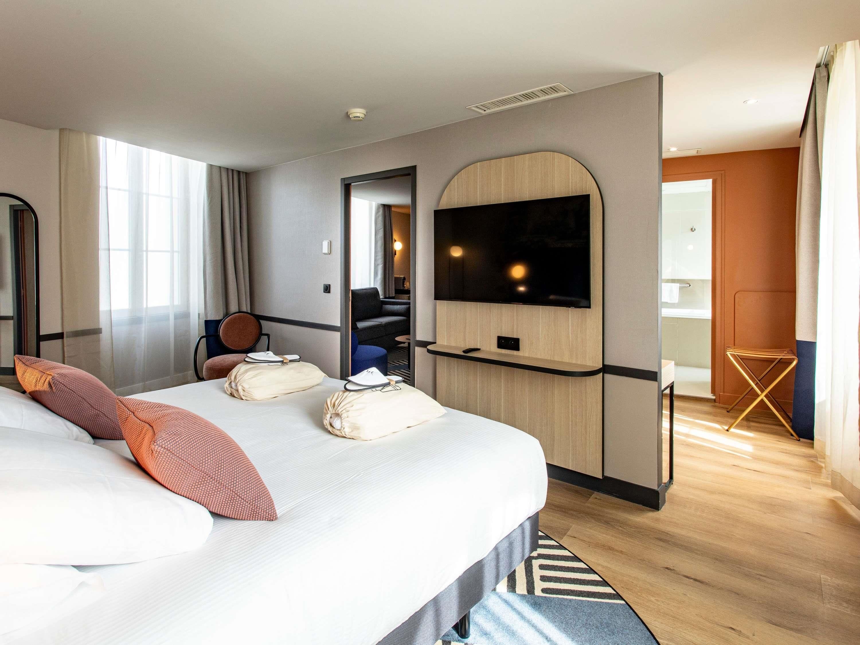 Hotellihuone, jossa on suuri sänky, seinälle asennettu taulutelevisio, pieni oleskelualue, jossa on tuoli ja pöytä, sekä kylpyhuone, joka näkyy avoimesta ovesta.
