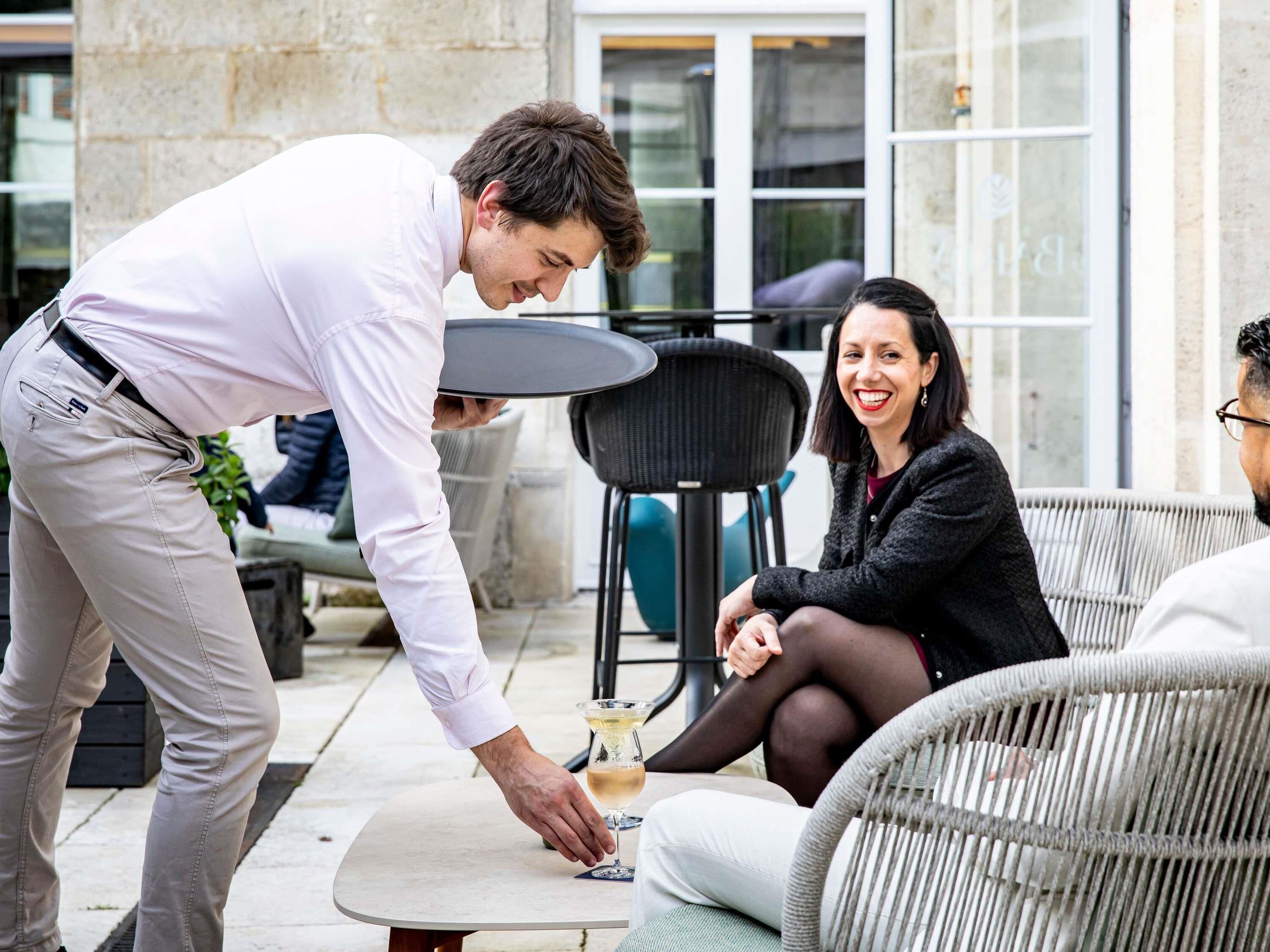 A waiter serving a glass of wine to a seated woman at an outdoor café with wicker furniture and a stone wall in the background.