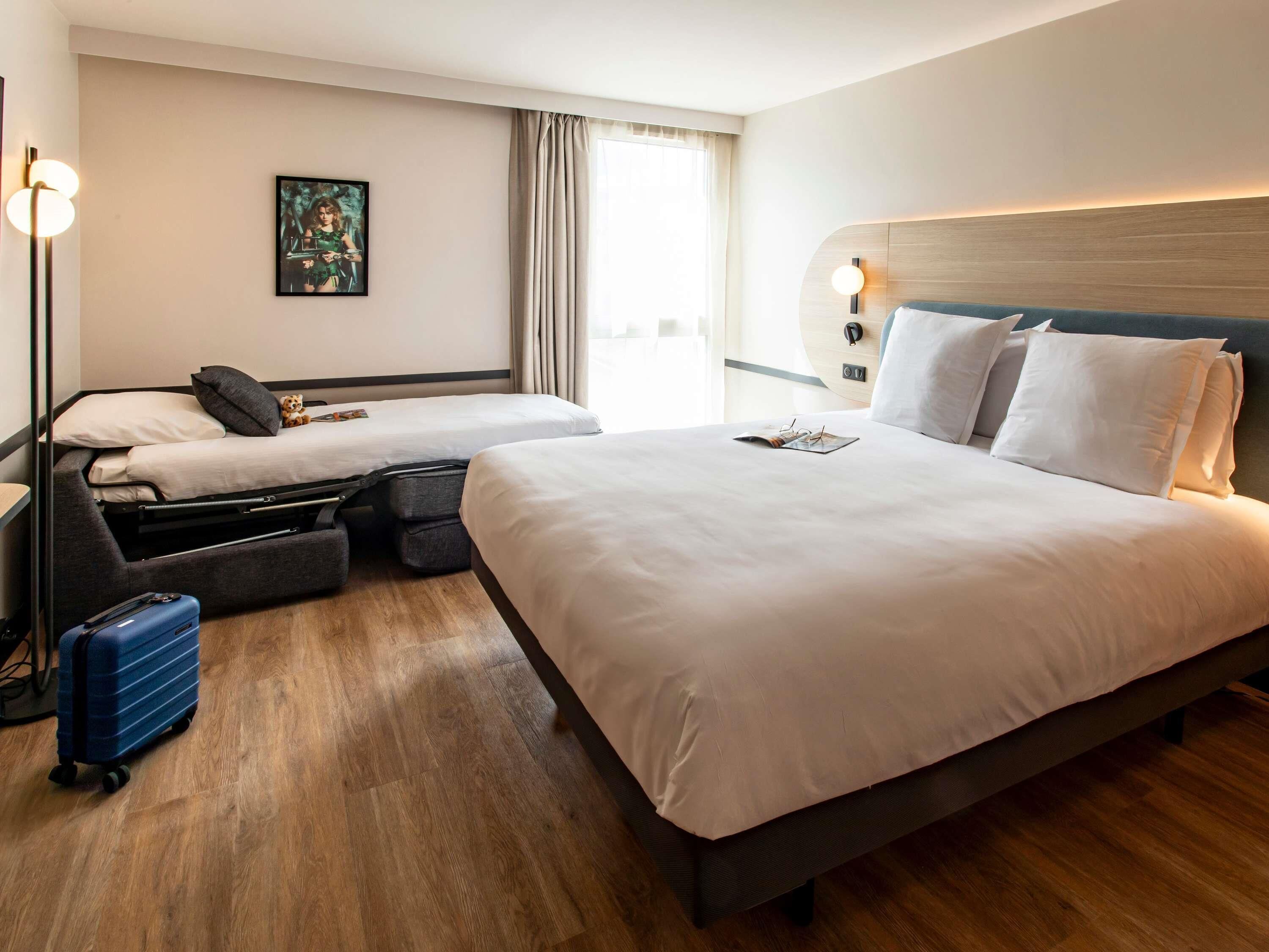 Habitación de hotel con dos camas, un escritorio, una silla, una maleta y un cuadro en la pared.
