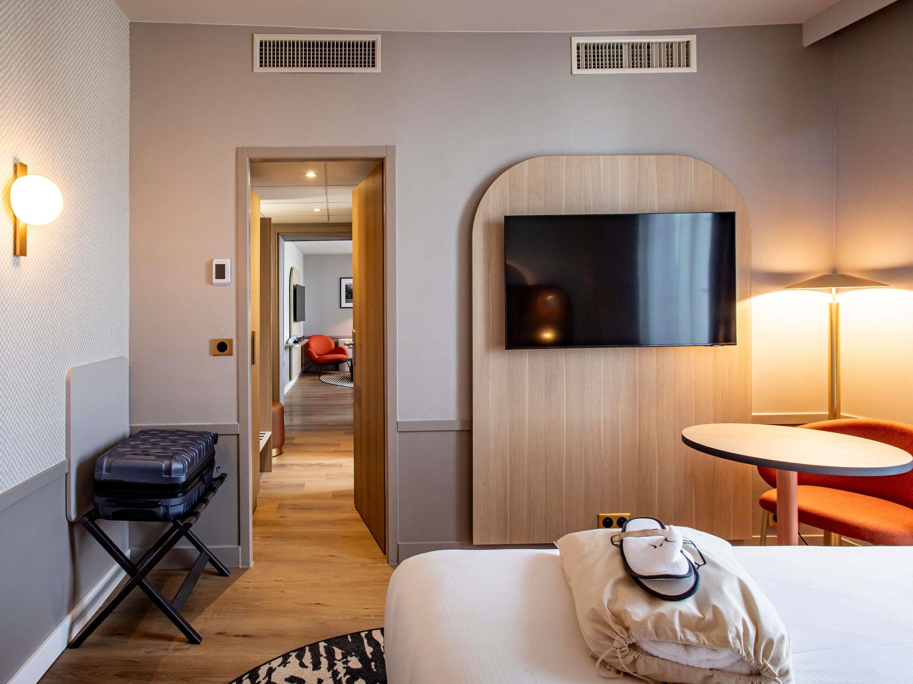 Une chambre d’hôtel avec un lit, un bureau, une chaise, un téléviseur fixé au mur et une valise posée sur le sol.