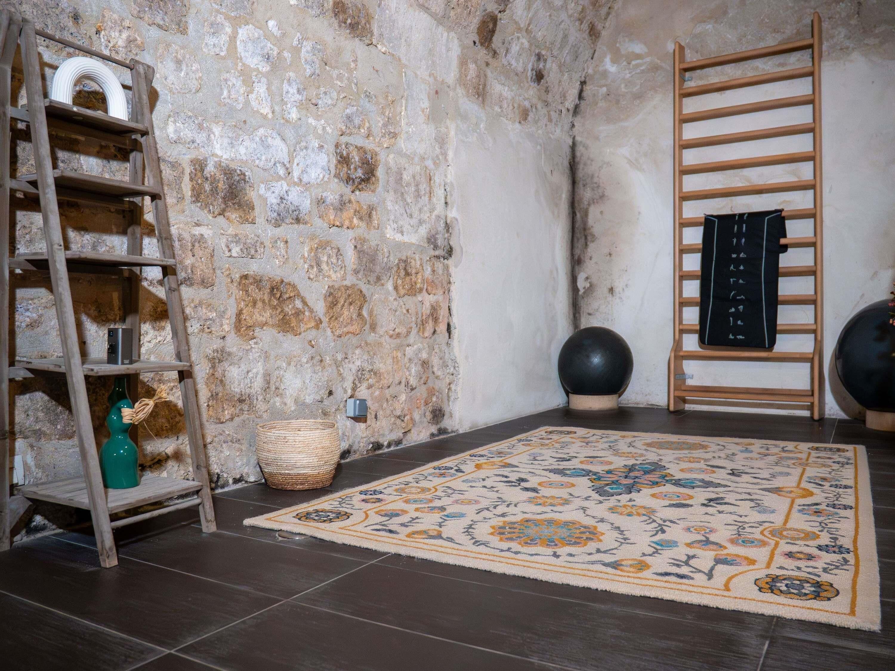 Una habitación rústica con pared de piedra, escalera de madera, una cesta tejida, alfombra estampada y un objeto esférico negro en el suelo.