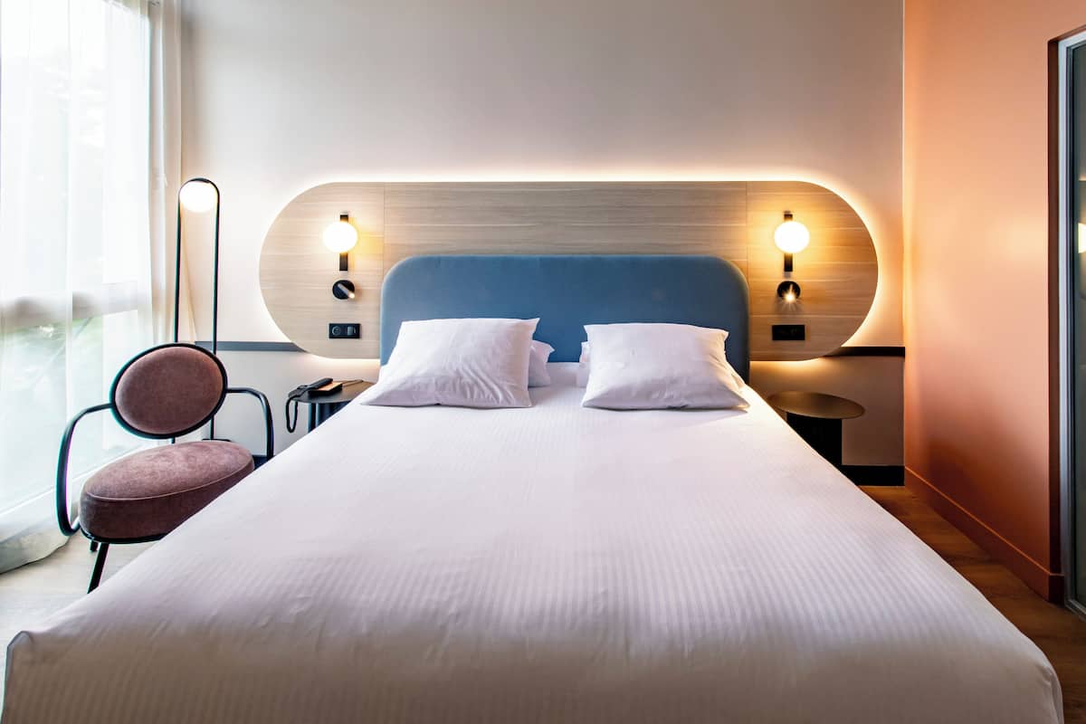Une chambre d’hôtel moderne avec un grand lit, deux tables de chevet, une chaise et une vue sur l’extérieur.