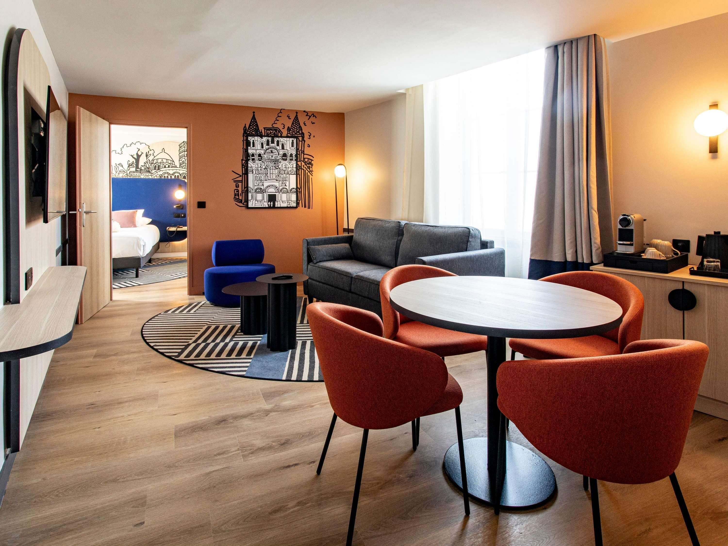 Ein modernes Hotelzimmer mit einem runden Esstisch, roten Stühlen, einem grauen Sofa, einem blauen Sessel und einem Bett mit Blick auf ein Gebäude.