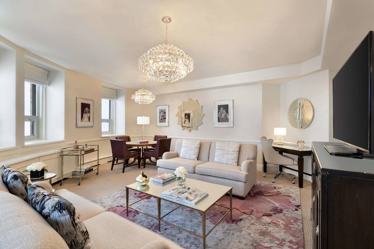 A modern living room with a sofa, coffee table, and a TV. There are two chandeliers, a dining table with chairs, and a desk with a lamp.