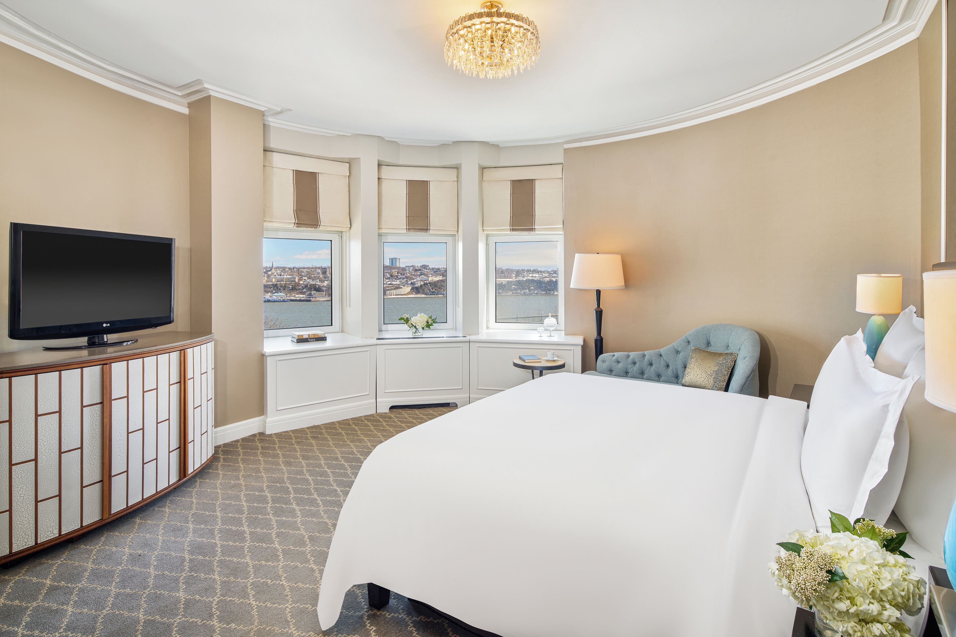 Quarto de hotel com uma cama grande, televisão, escrivaninha com abajur e janelas com vista para a cidade.