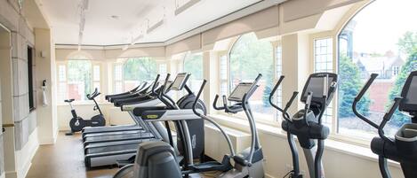 Un gimnasio con numerosas caminadoras y bicicletas estáticas, amplias ventanas que ofrecen vistas a edificios y árboles, y un diseño moderno y cuidado.