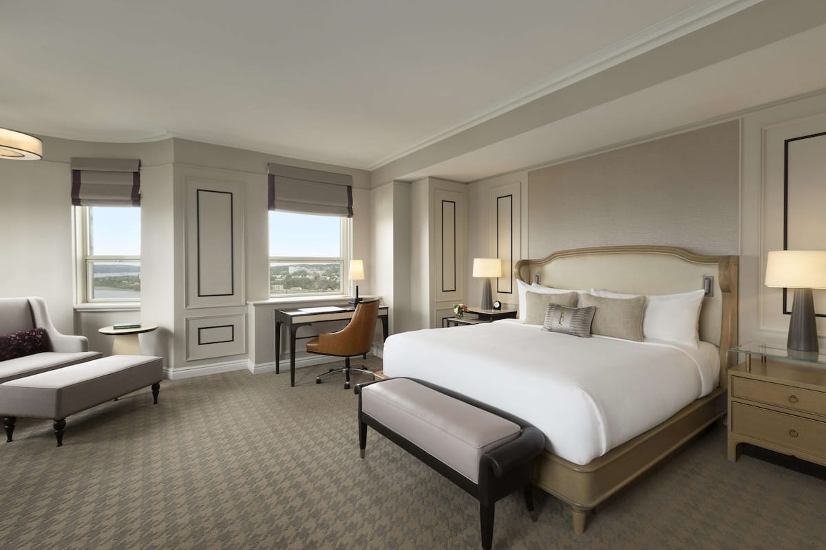 A hotel room with a large bed, a desk, a chair, a bench, and a nightstand with a lamp.