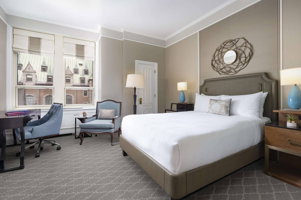A hotel room with a large bed, a desk with a chair, a small table with a lamp, and a window with a view of buildings.