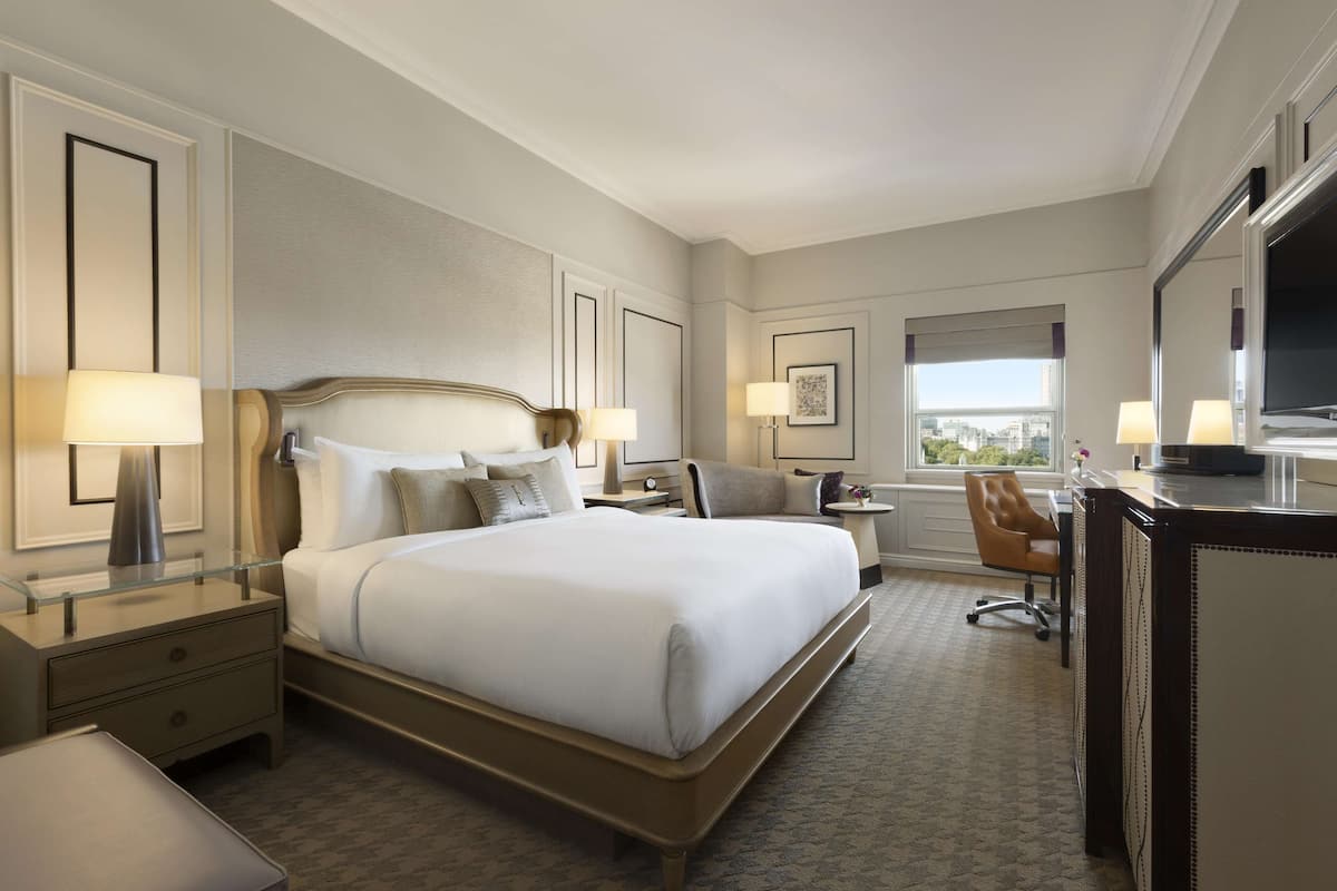 A hotel room with a large bed, a desk, a chair, a TV, and a window with a city view.