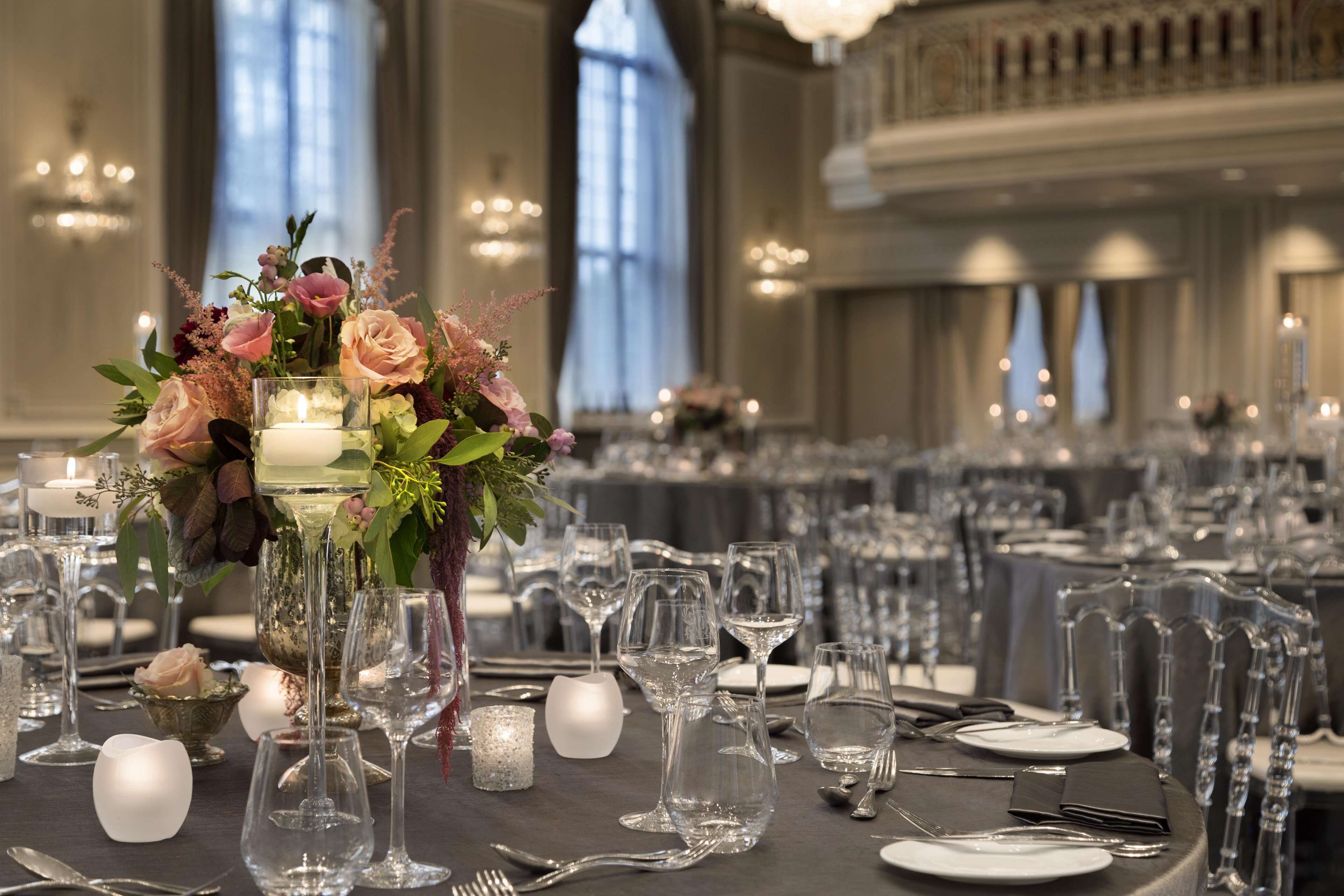 Un salón de banquetes con mesas dispuestas para un evento formal, con centros de mesa florales, velas y copas de cristal.
