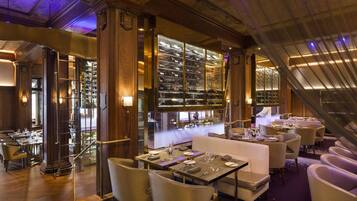 Une salle à manger luxueuse, dotée de hauts plafonds, de boiseries décoratives et d’une grande cave à vin visible à travers une paroi de verre.