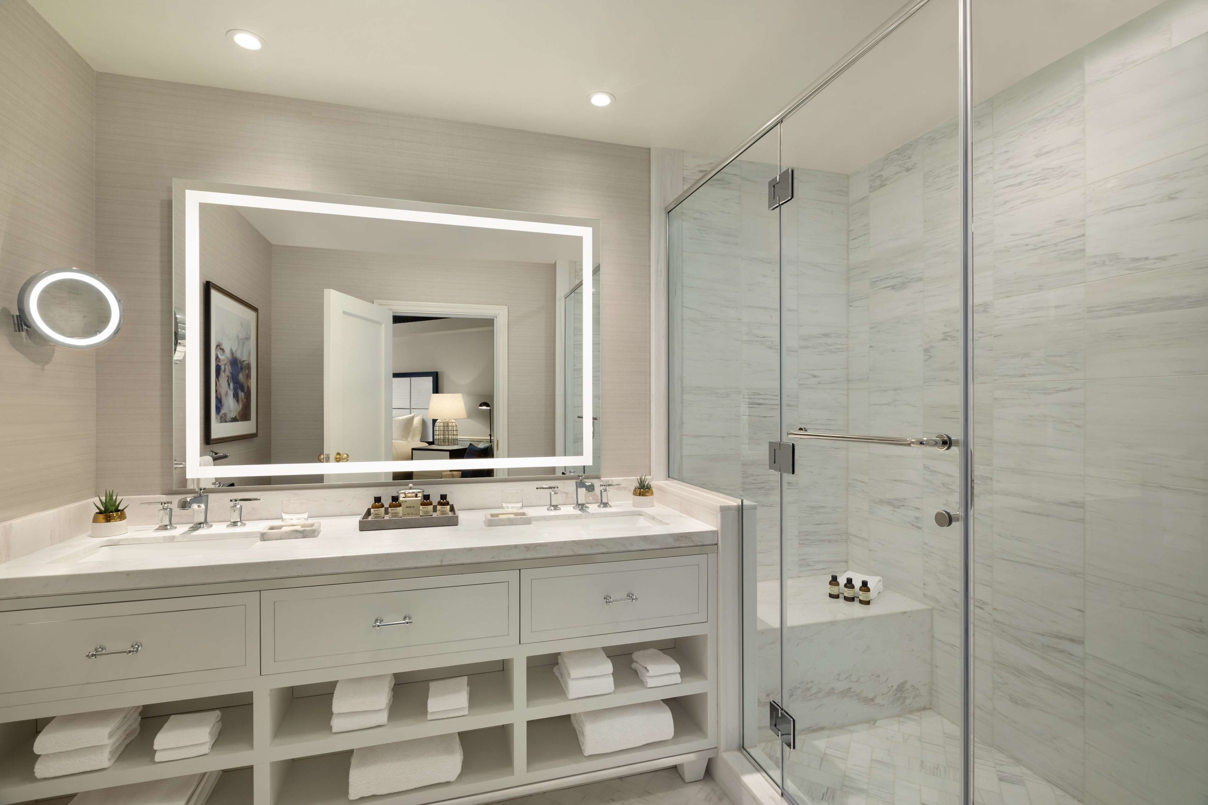 A modern bathroom with a large mirror, two sinks, a shower, and a vanity with storage.