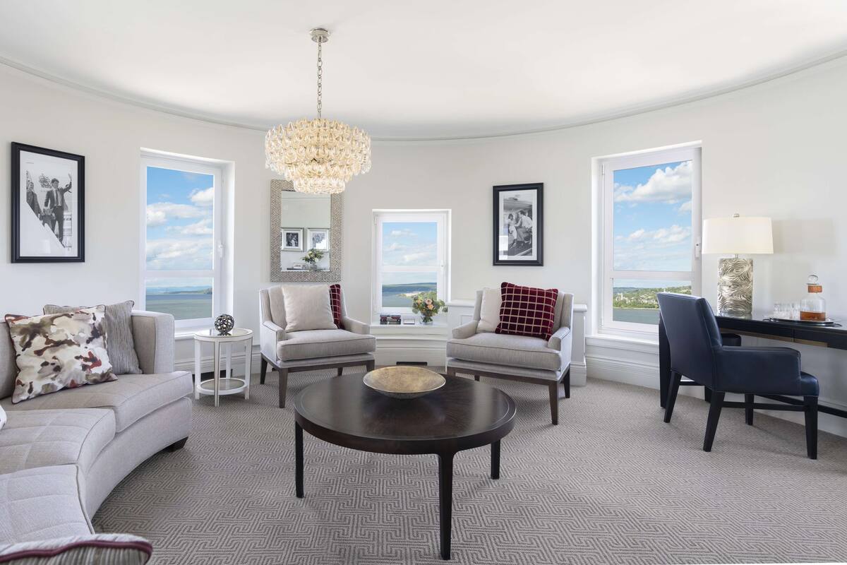 A modern living room with a round coffee table, two armchairs, a sofa, and a dining table. There are large windows with a view of the ocean, a chandelier, and framed pictures on the walls.