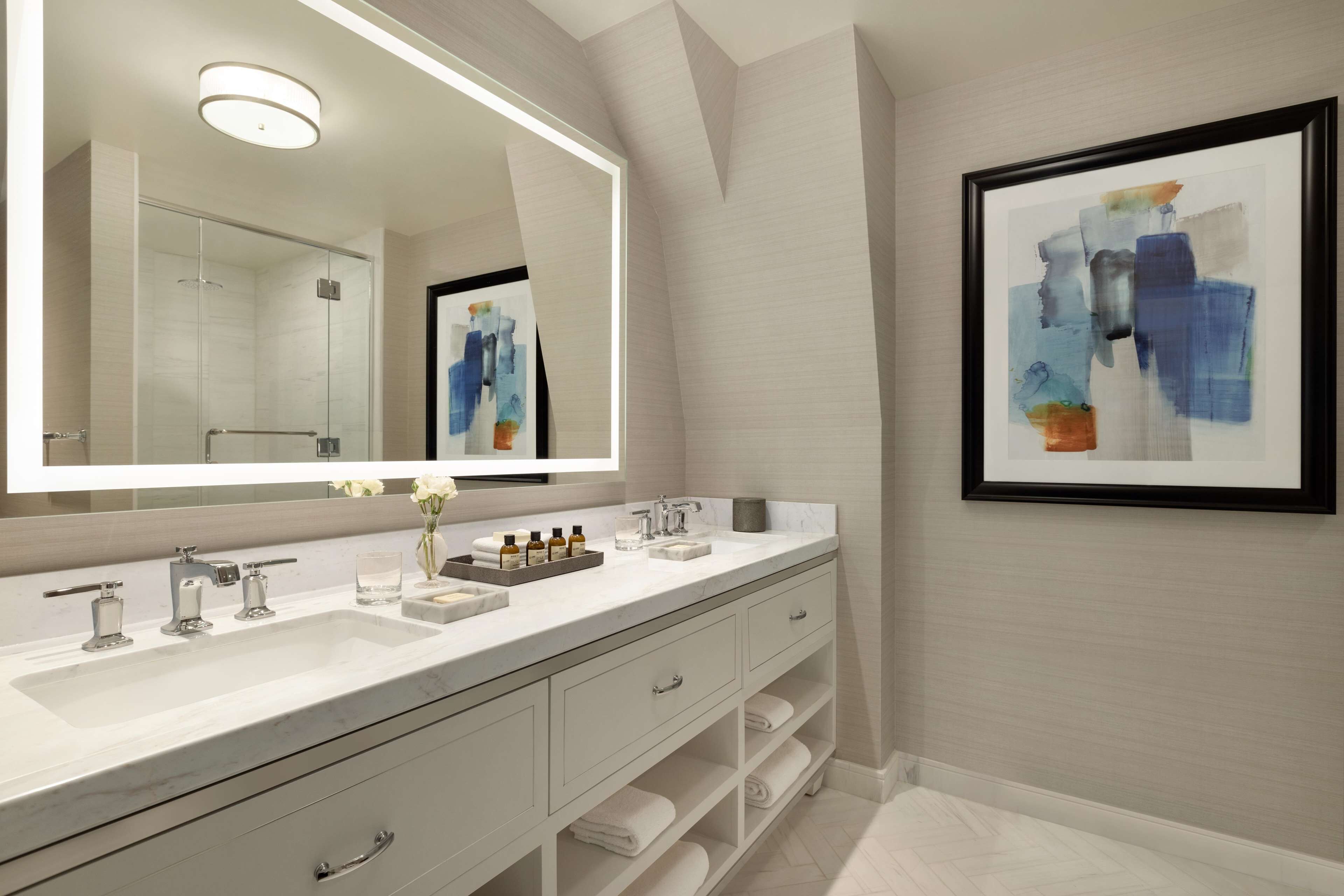 A modern bathroom with a large mirror, two sinks, a shower, and a framed abstract painting on the wall.