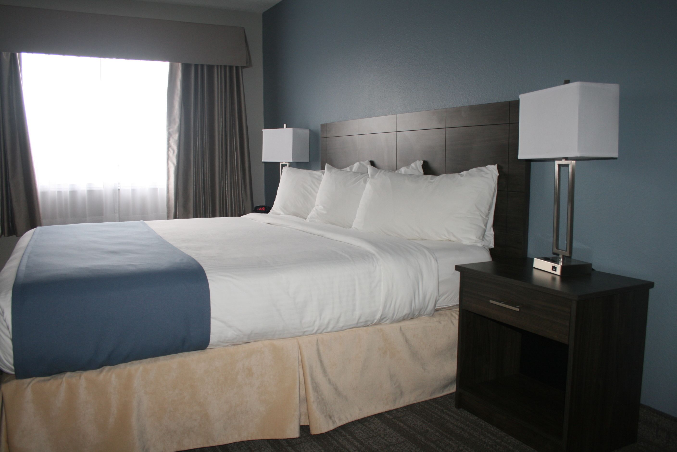 A hotel room with a large bed, two bedside tables with lamps, a window with curtains, and a dark-colored headboard.