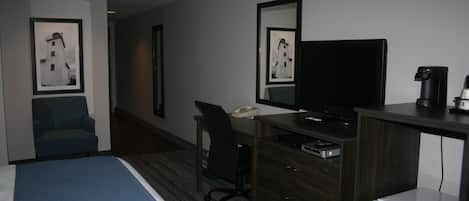 A hotel room with a bed, a desk with a computer, a chair, a small refrigerator, and two framed pictures on the wall.