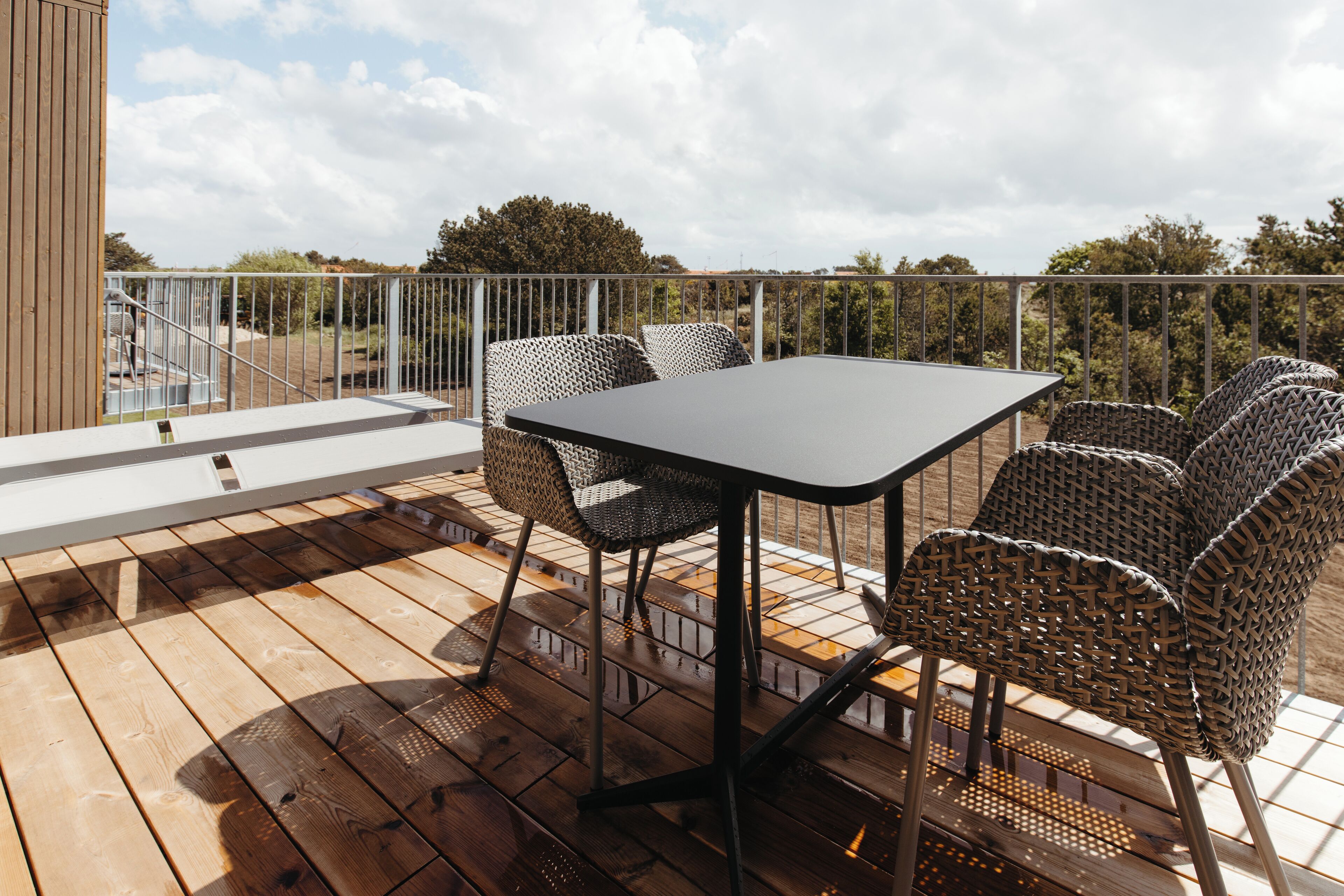 A wooden deck with a table and wicker chairs, overlooking a fenced area with trees.