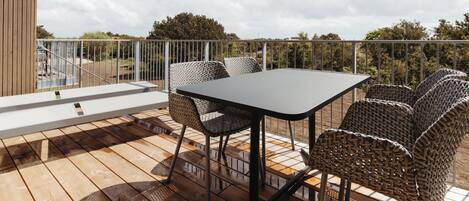 A wooden deck with a table and wicker chairs, overlooking a fenced area with trees.