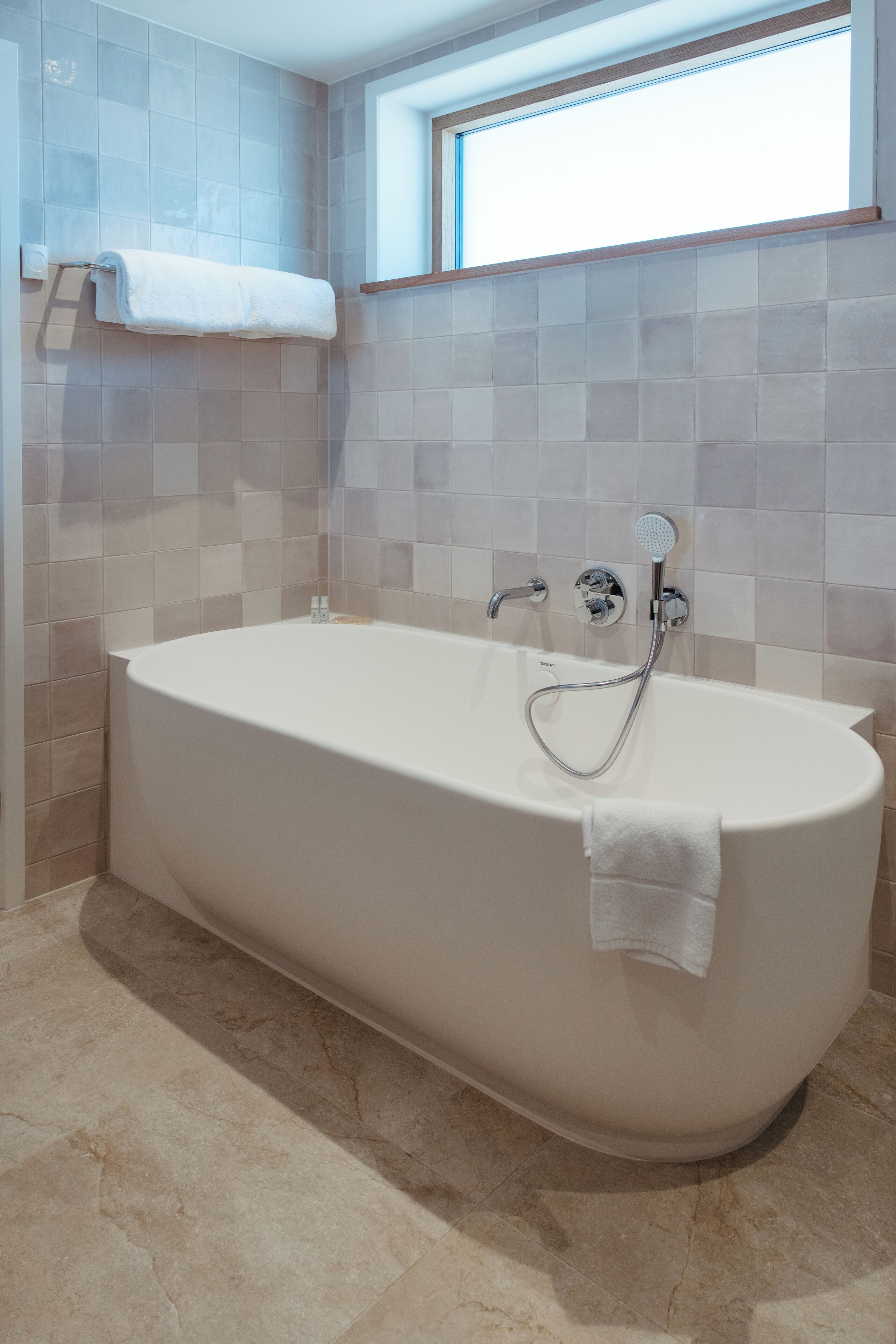 Un baño moderno con una bañera independiente, paredes con azulejos y una ventana sobre la bañera.