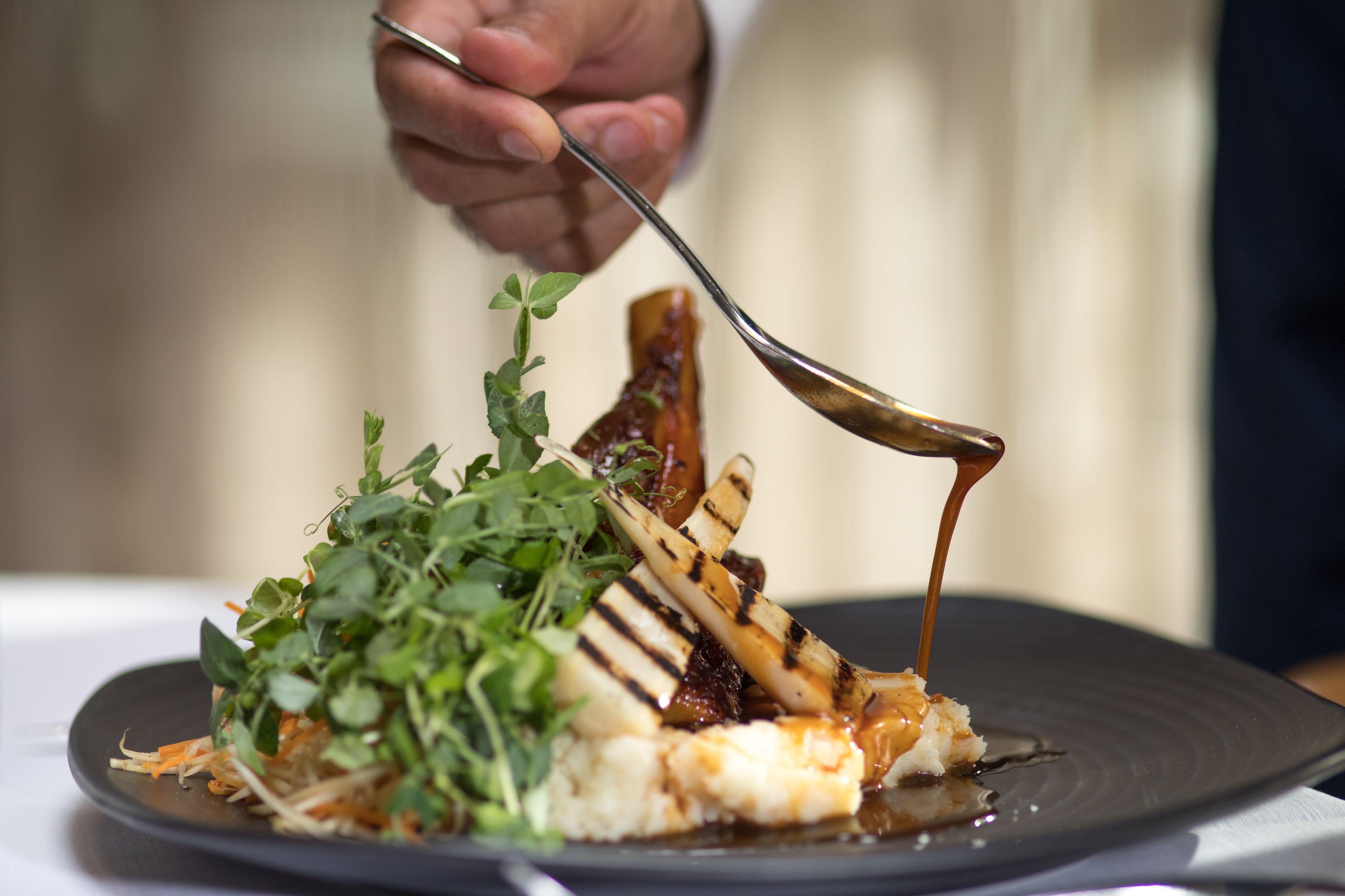 A person serving a dish with grilled meat, greens, and sauce on a plate.