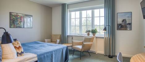 A hotel room with a bed, a desk, a chair, a window with curtains, a radiator, and a plant.