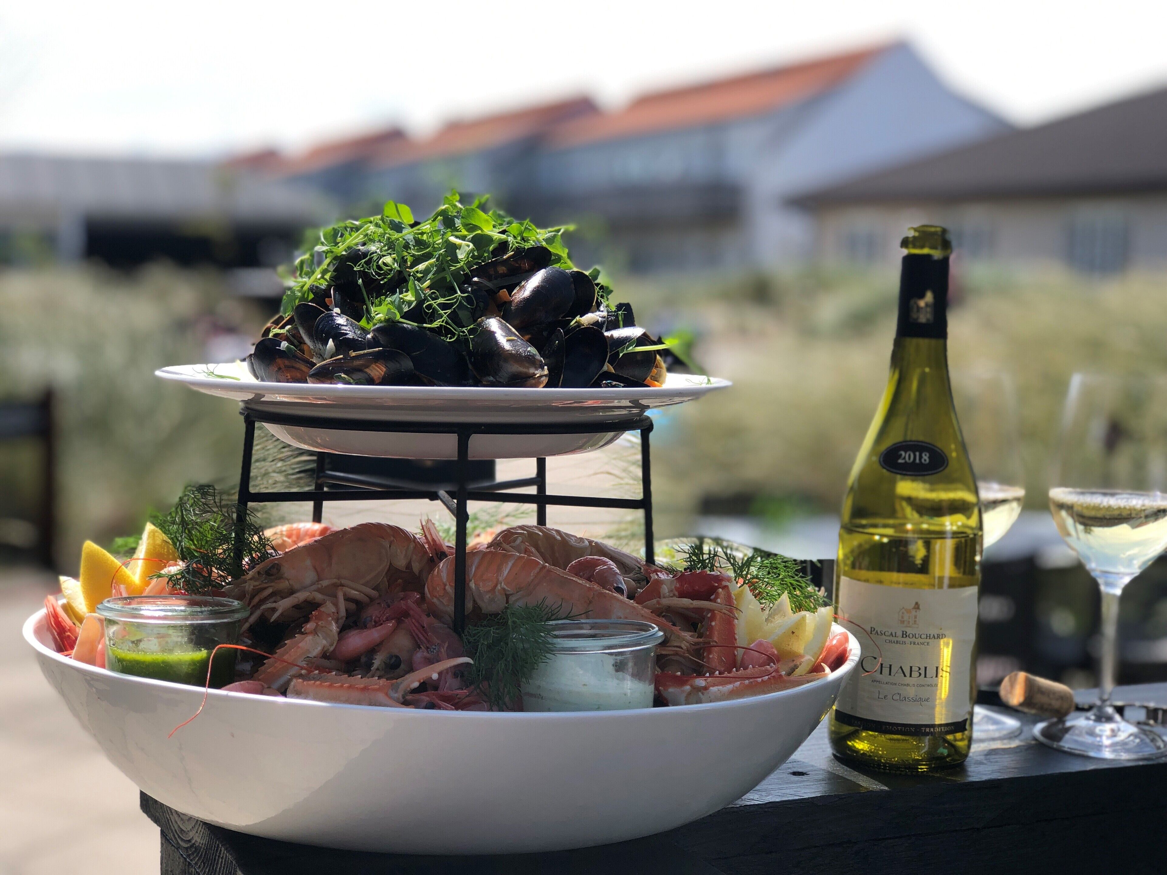 Un plateau de fruits de mer avec des moules et des crevettes, une bouteille de vin et des verres sur une table à l’extérieur.