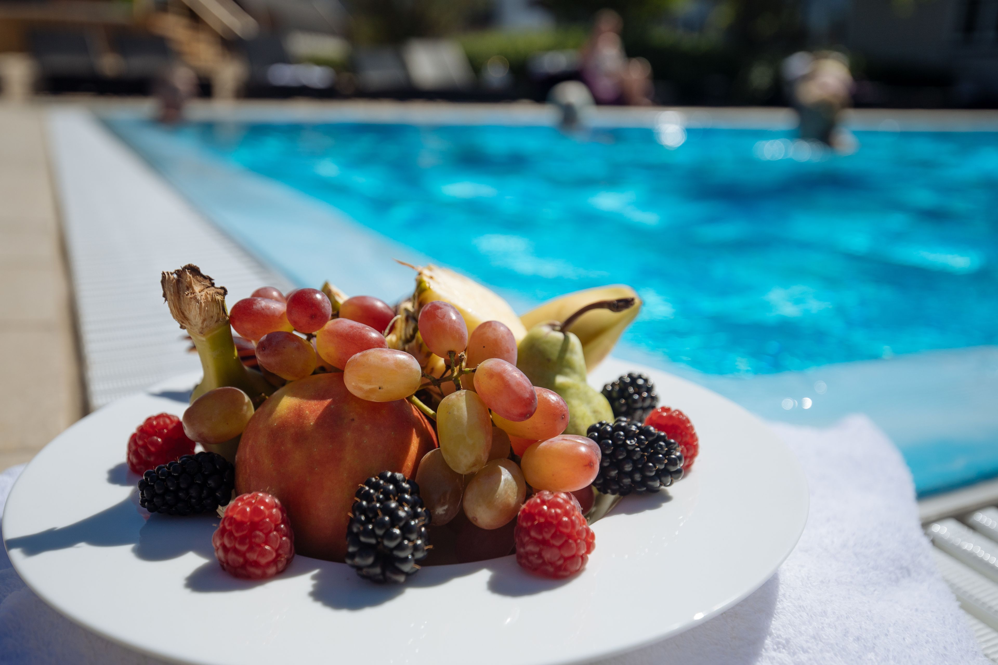 Uma travessa de frutas sobre um prato branco, perto de uma piscina com vista clara para a área da piscina.